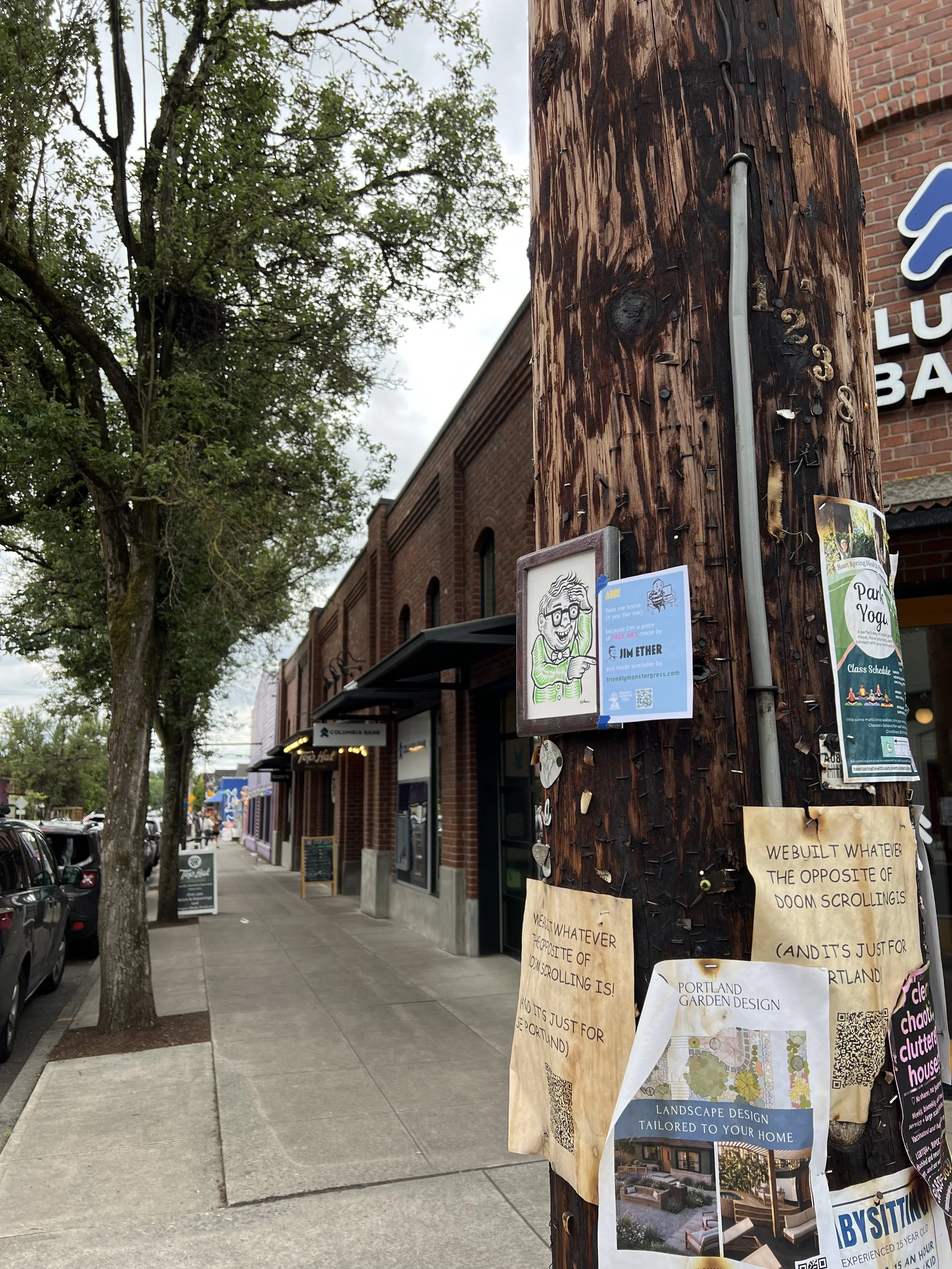 A sidewalk scene in a small town with a utility pole covered in flyers and notes, including one that says "WE BUILT WHATEVER THE OPPOSITE OF DOOM SCROLLINGS!". There are trees, storefronts, and parked cars along the street.