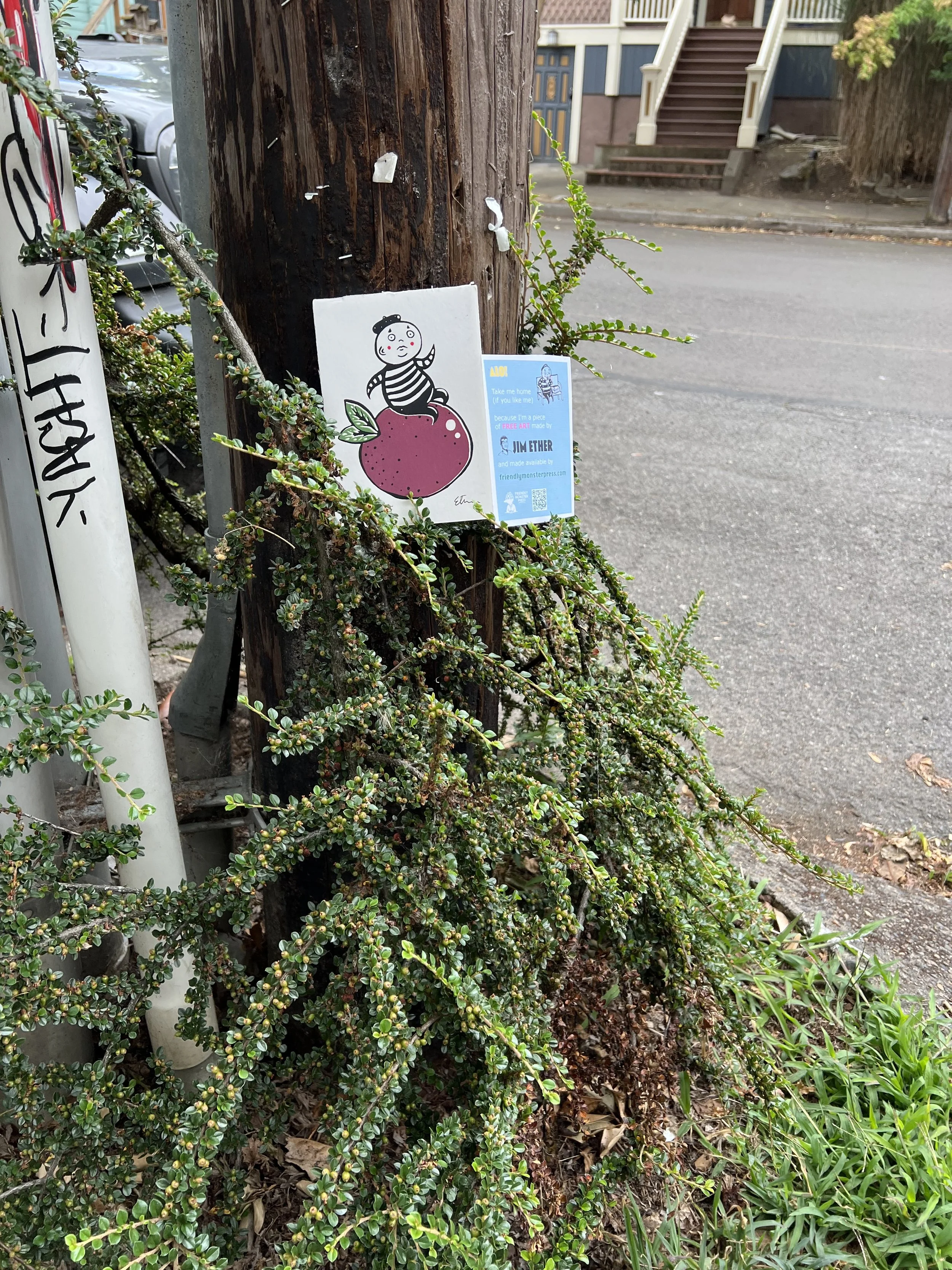 A wooden utility pole with attached stickers and a sign, surrounded by green shrubbery and plants. A residential street with steps and houses is visible in the background.