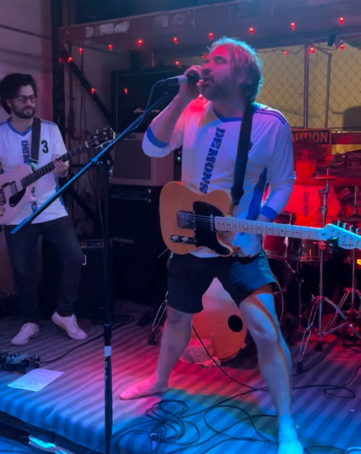 A band performing on stage with one member singing into a microphone and playing an electric guitar, another member playing an acoustic guitar in the background, under colorful stage lighting.