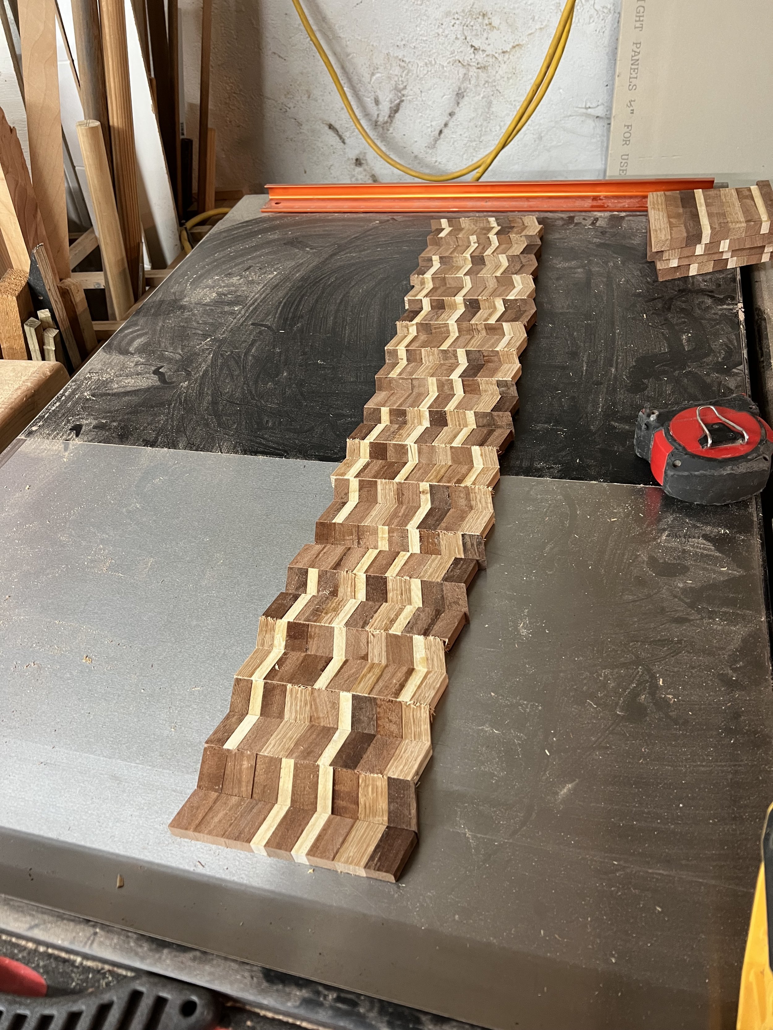 A long row of small wooden hexagon-shaped pieces on a black workbench in a woodworking shop, with tools and supplies around.