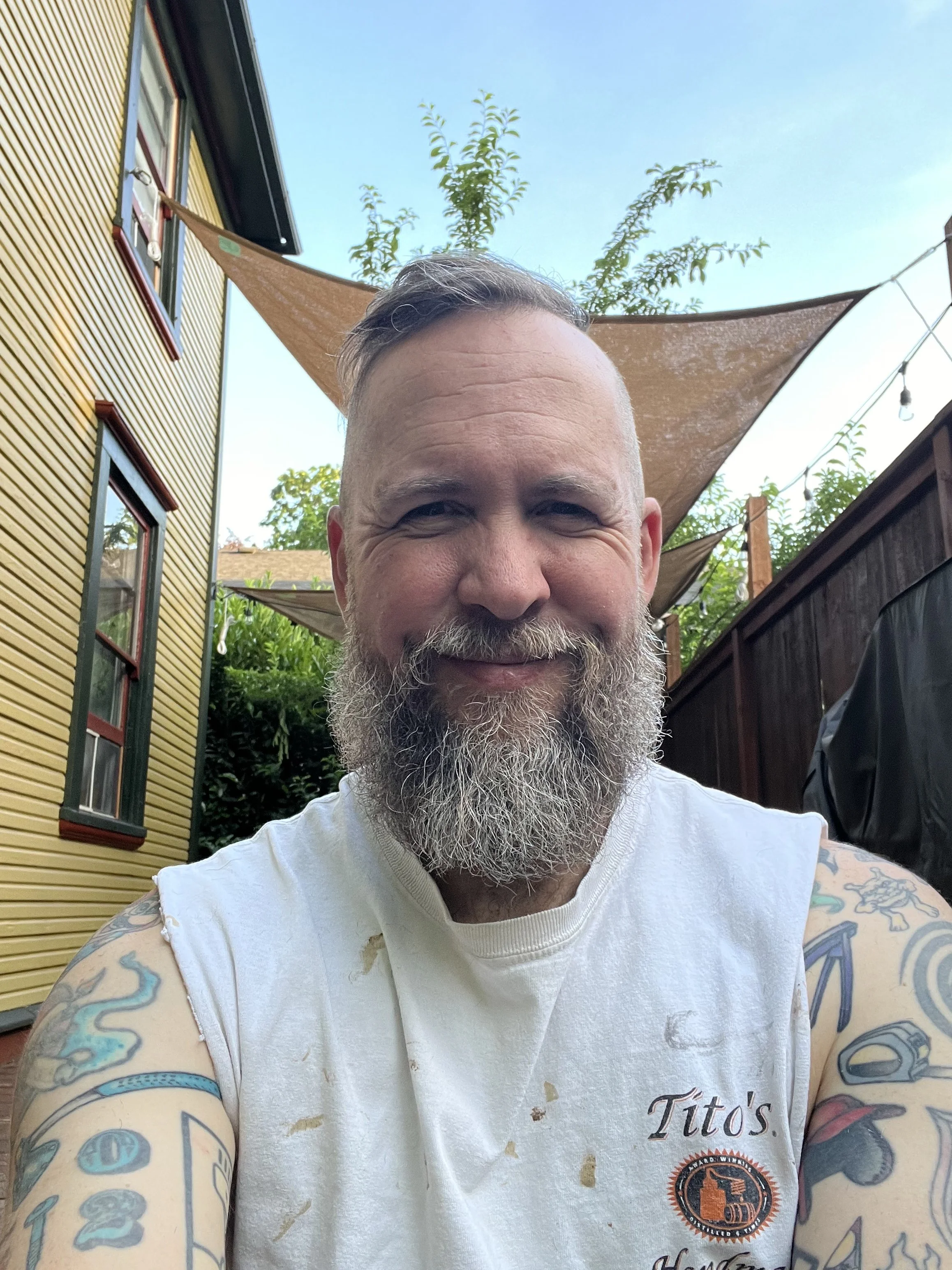 A smiling man with a beard and tattoos on his arms, wearing a white sleeveless Titos shirt, taking a selfie outdoors. There are yellow and brown buildings, trees, and a blue sky in the background.