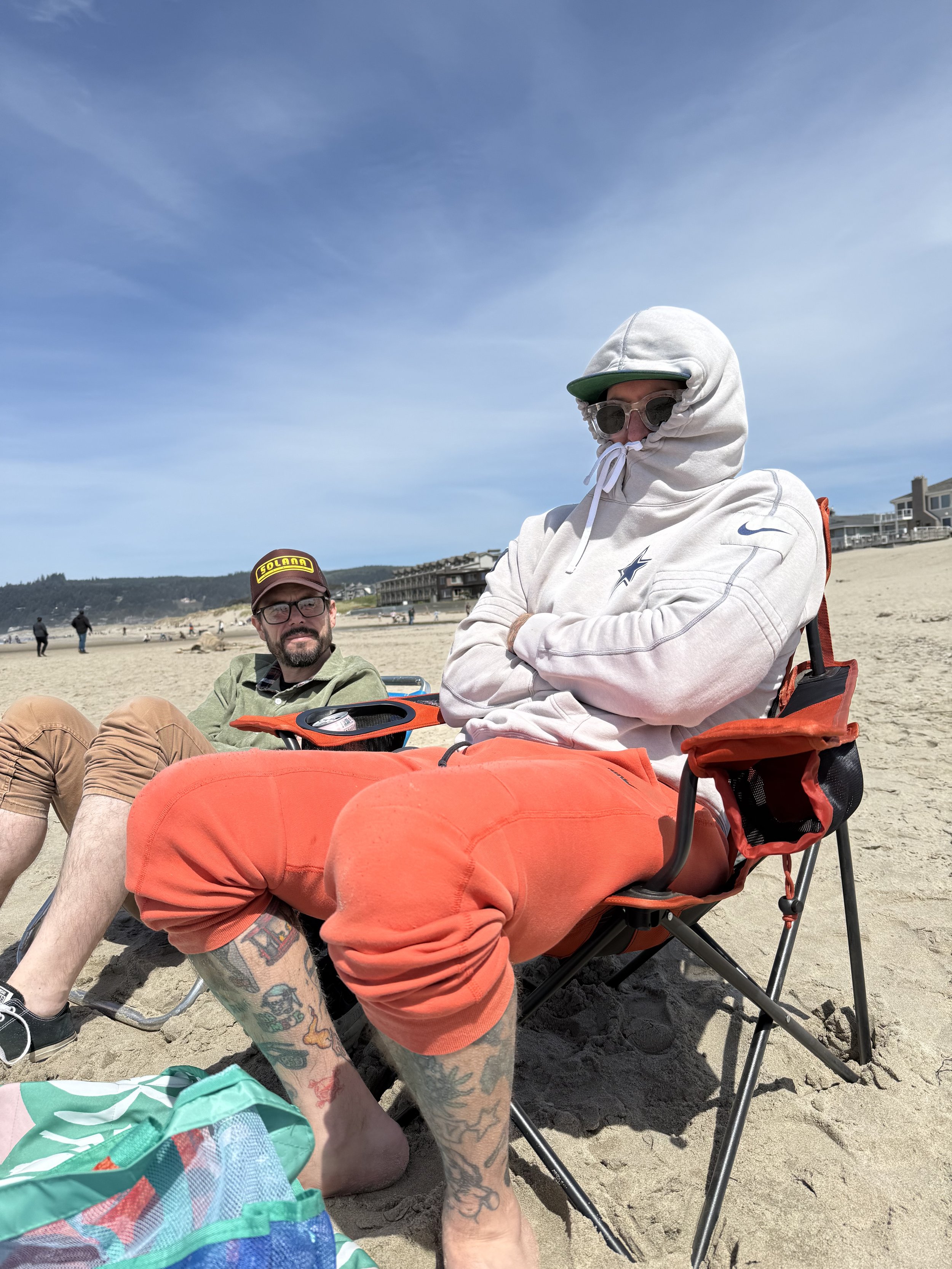 Two men sitting on a beach with sand and a blue sky, one wearing a white hoodie and sunglasses with arms crossed, the other wearing a cap, glasses, and a green hoodie. There is a backpack, a foldable chair, and beach chairs in the background.