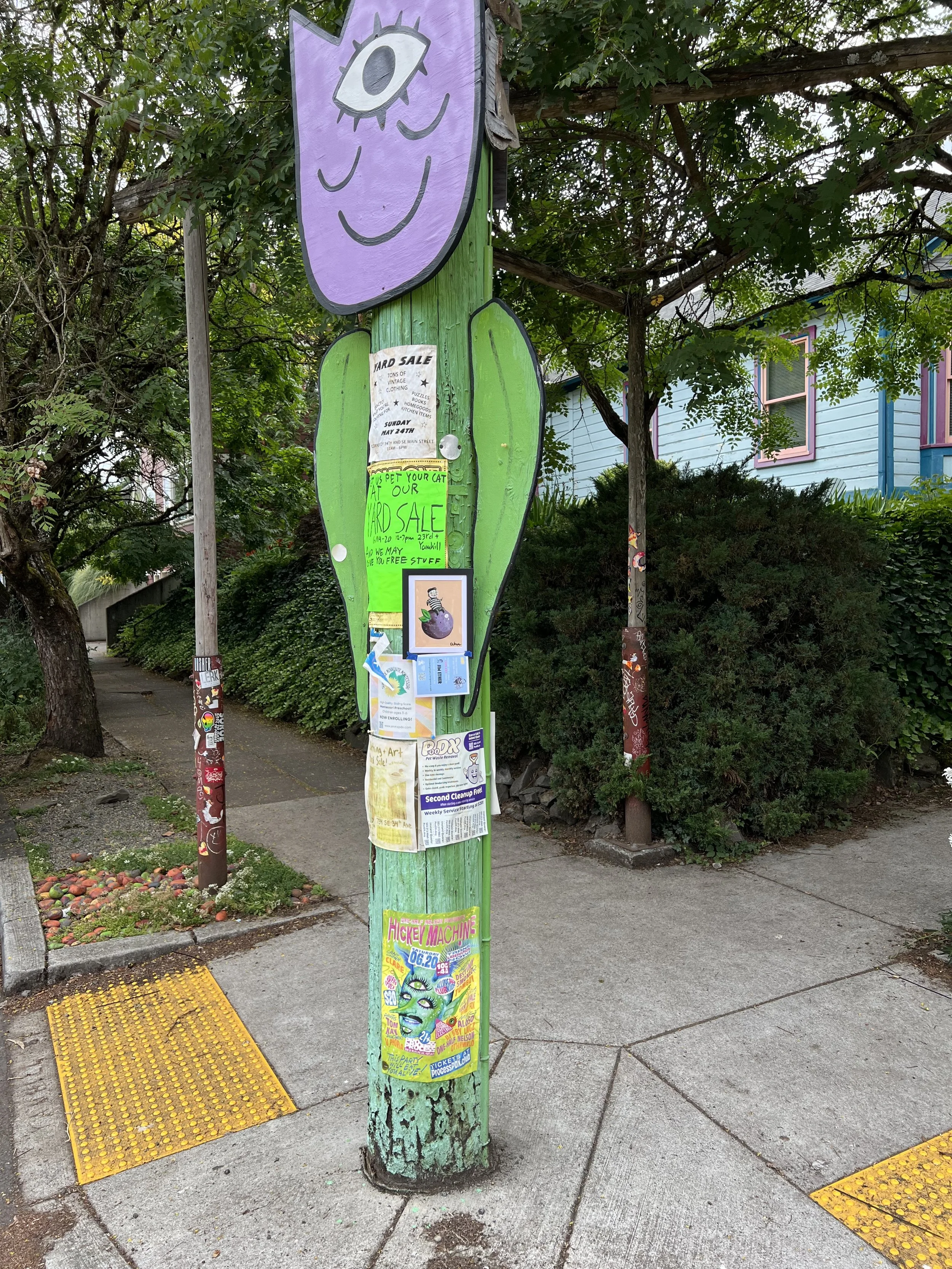 Colorful sidewalk pole with various posters, stickers, and artwork in a park-like setting with trees and residential houses in the background.