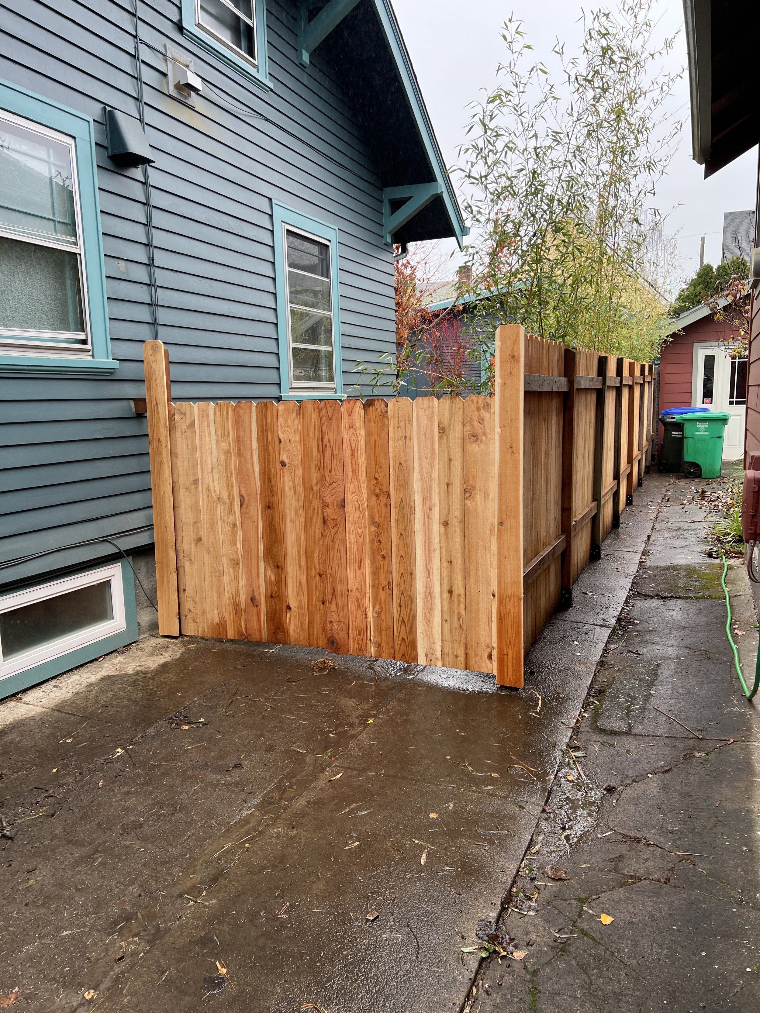 A newly built wooden fence runs along the alley, and there are bamboo trees with autumn leaves. Two wheelie trash bins are visible near a red house at the end of the alley, with a wet asphalt ground that has some leaves. Scott Cannon built that fence