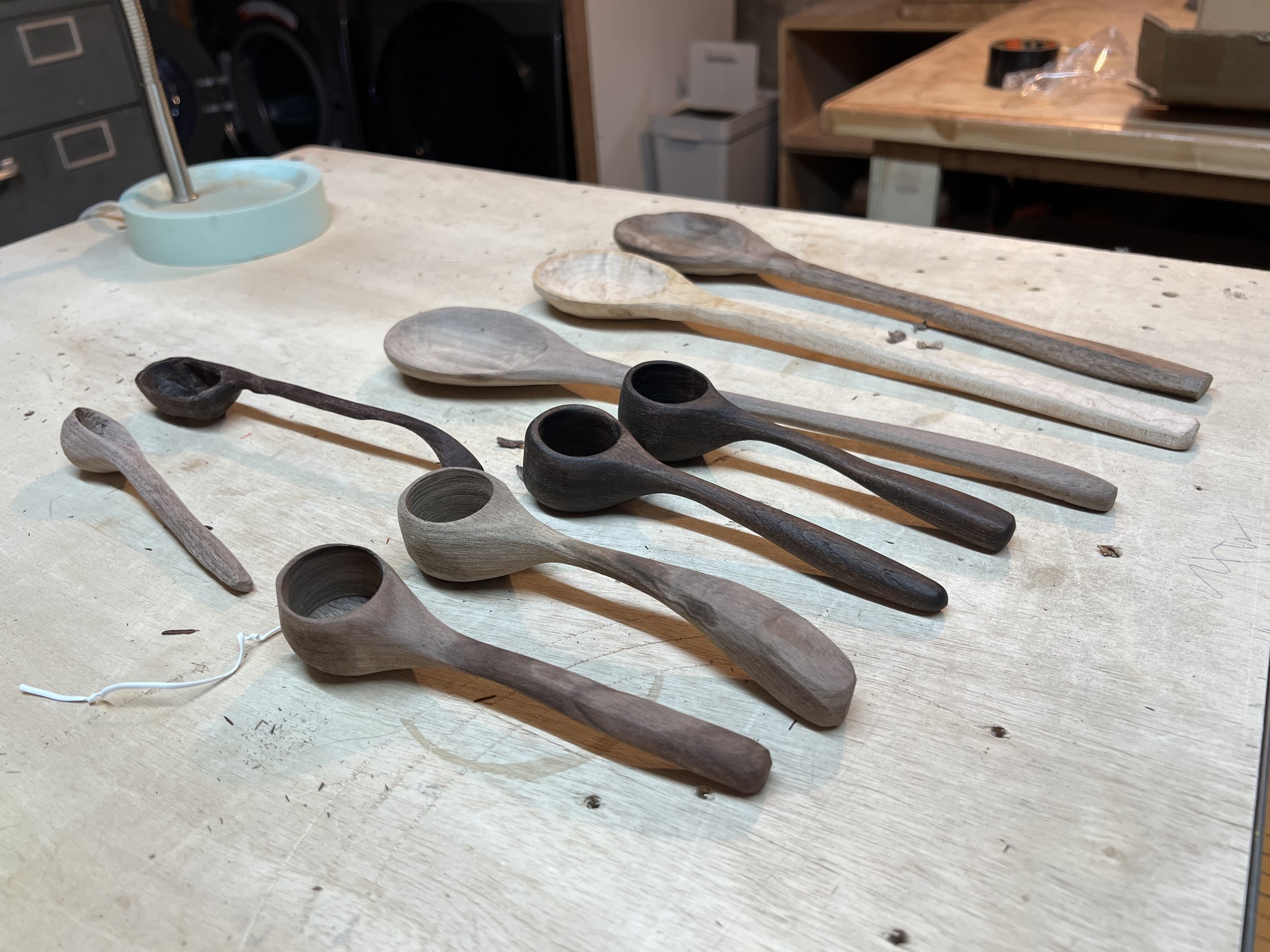 Various wooden spoon protoytpes are arranged on a light-colored wooden work surface in a workshop. The background shows woodworking tools and furniture.
