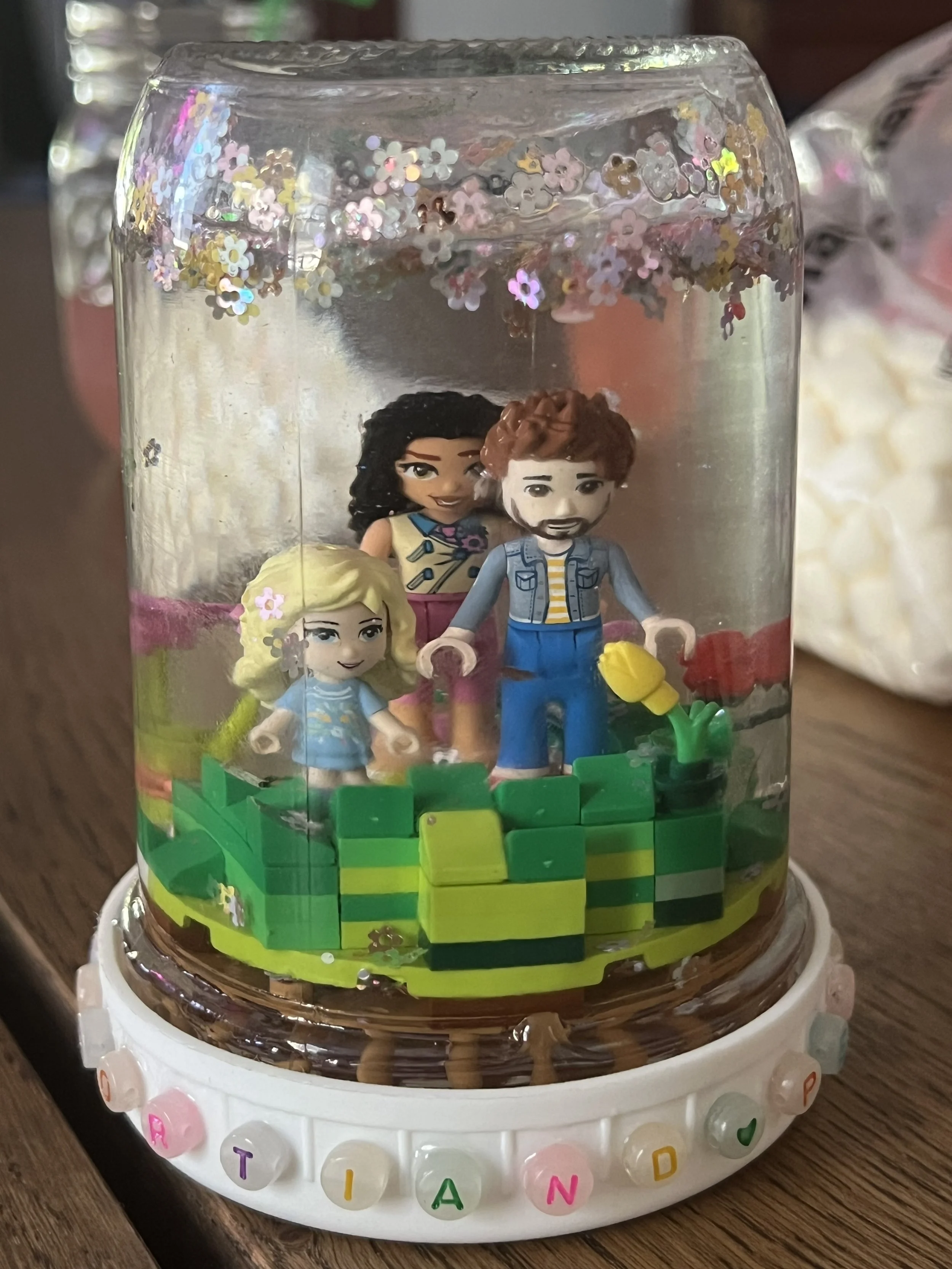 A transparent snow globe with small figurines of three children and colorful flowers inside, sitting on a wooden surface, with lettered beads around its white base.