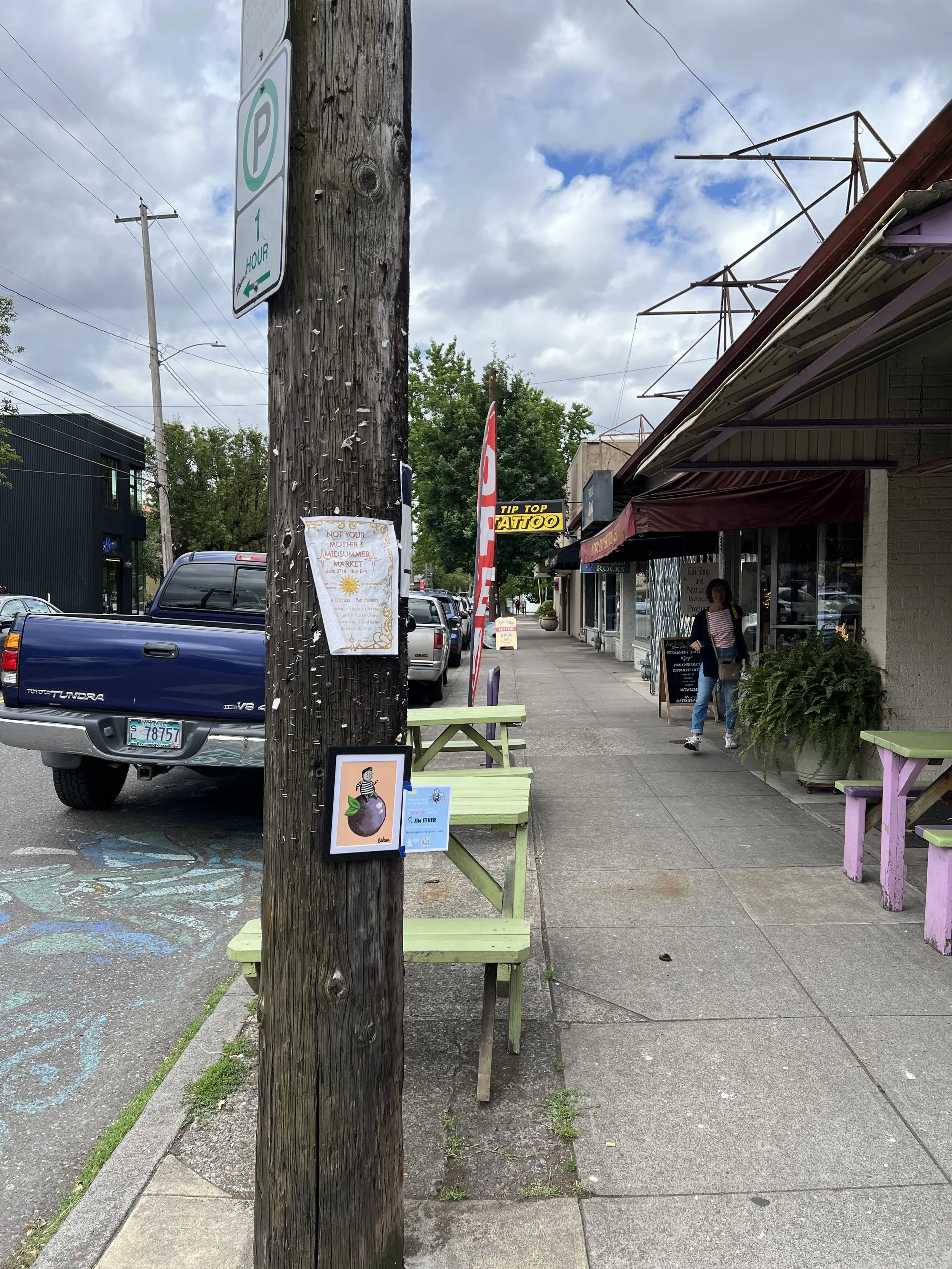 A street view with parked cars, a utility pole, storefronts with awnings, signs including a tattoo sign, benches, and a woman walking on the sidewalk under a partly cloudy sky.