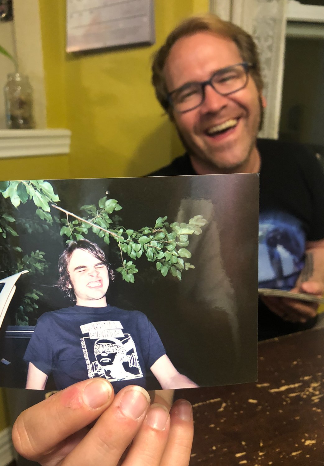 A person holding a photo of a young man with dark hair and a graphic t-shirt. The person in the background has glasses and is smiling.