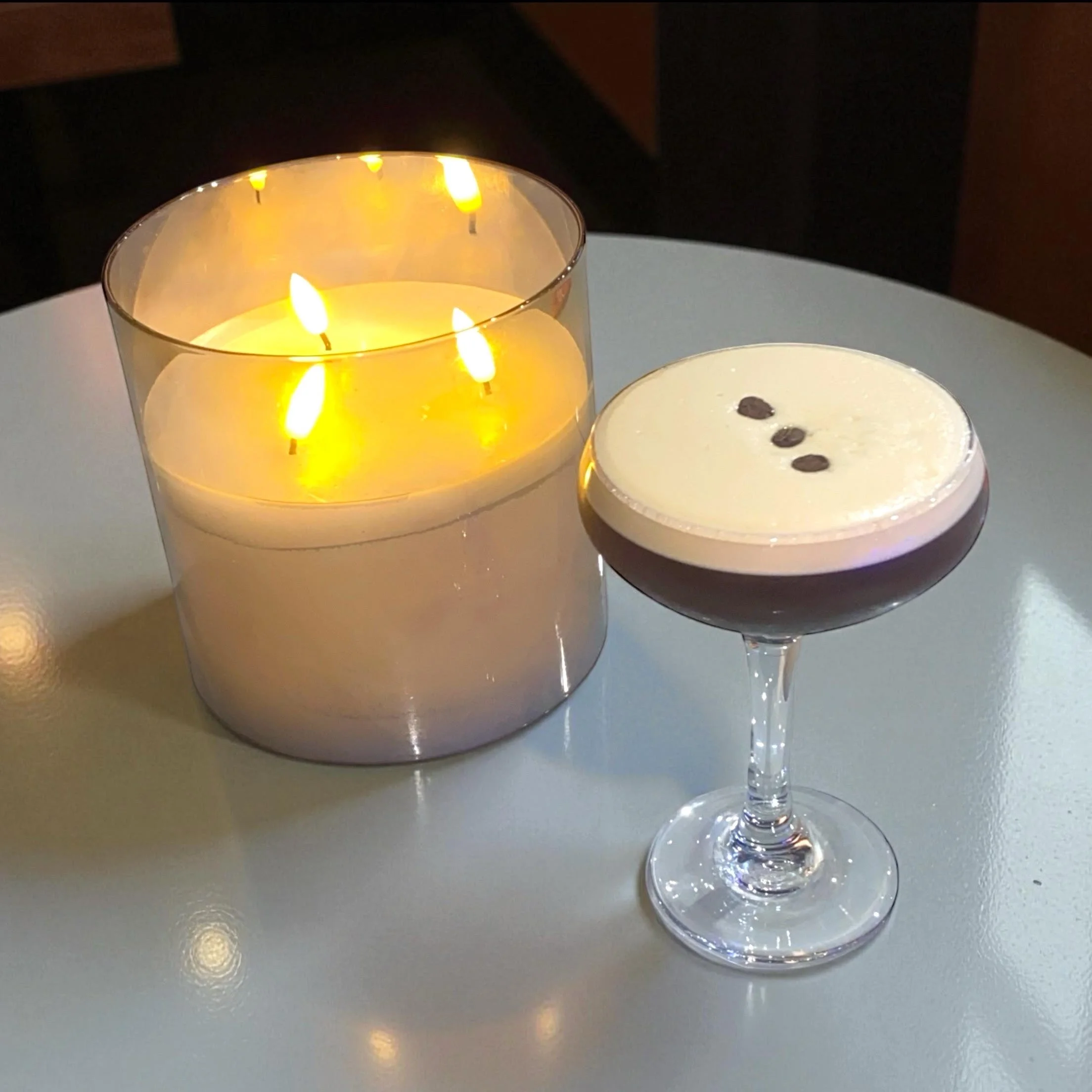 A lit candle in a glass holder and a dark cocktail with a frothy top topped with three coffee beans, on a white table.