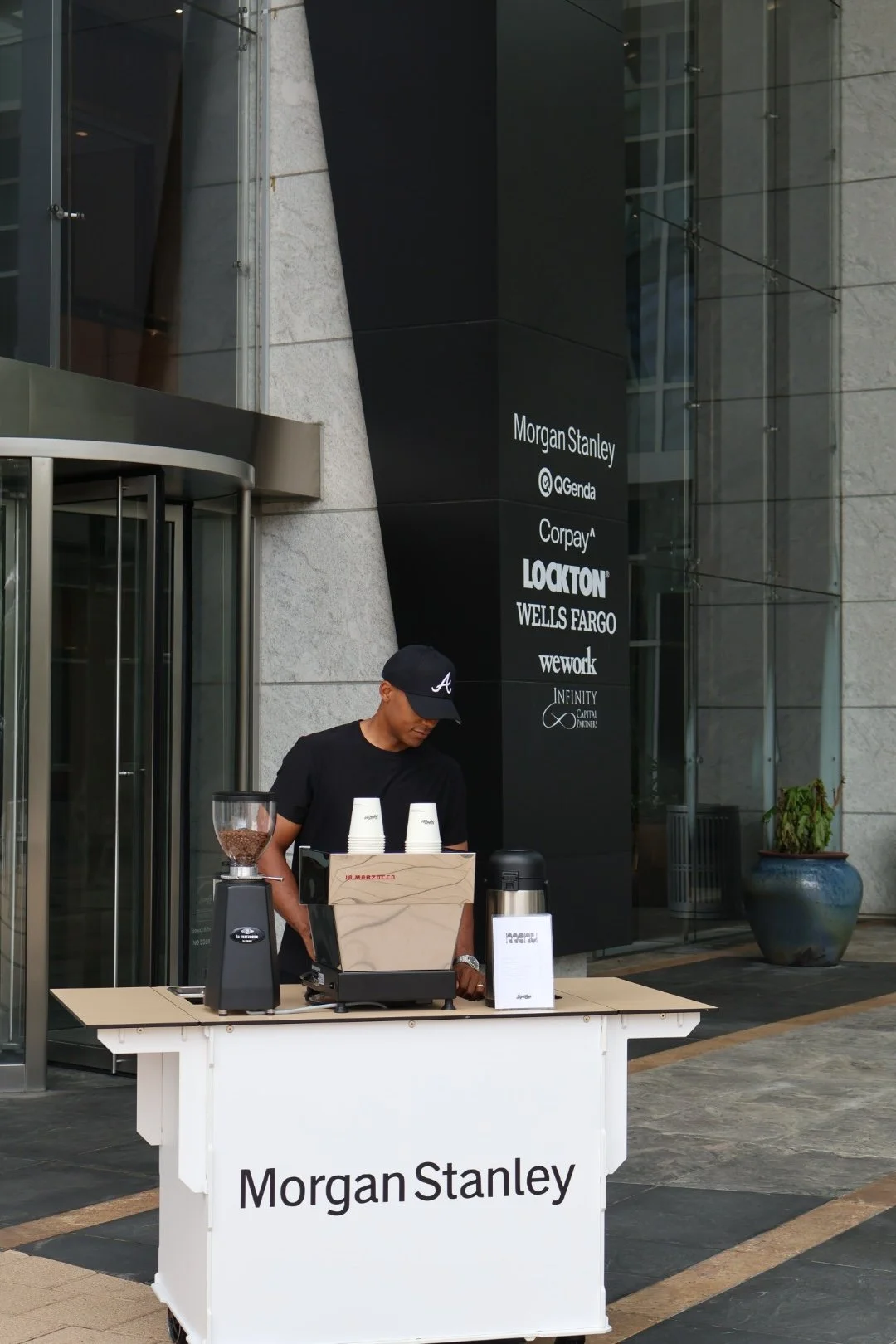 A man stands at a Morgans Stanely branded coffee cart outside a modern glass and concrete building, preparing a beverage with coffee beans and cups on the counter.