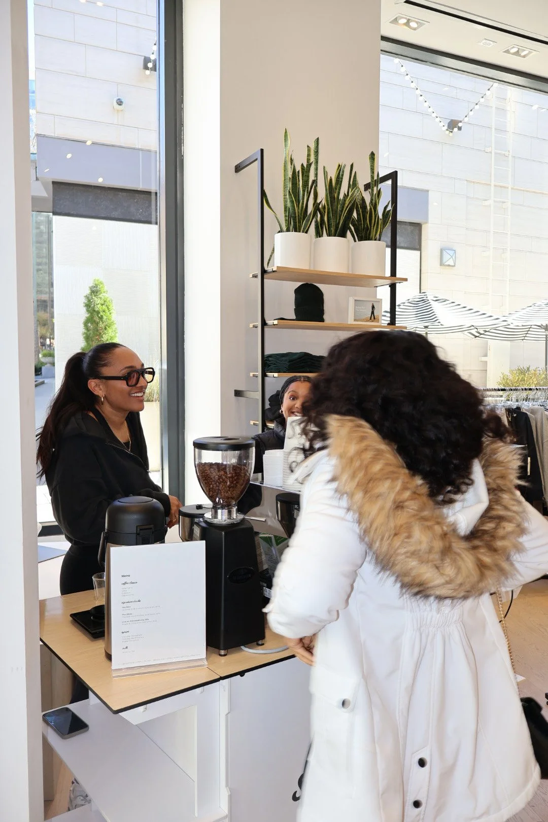 A woman in a white coat standing at a coffee shop counter, interacting with a barista, with another woman smiling behind the counter.