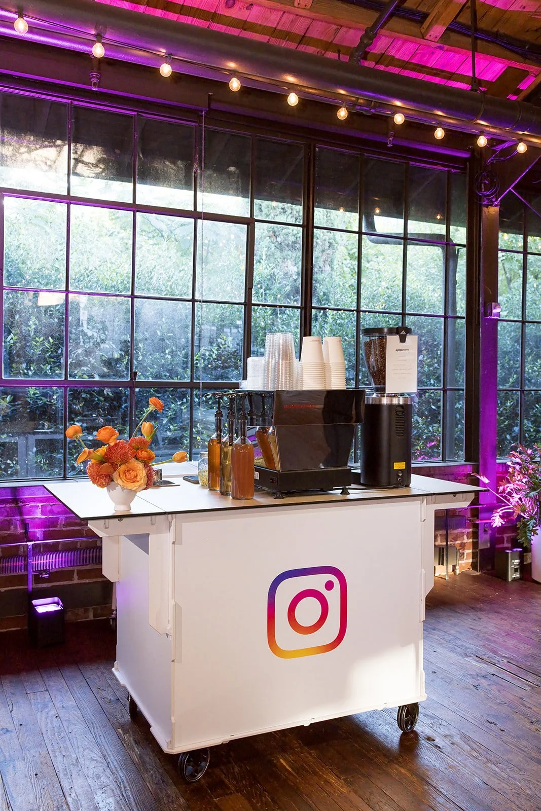 A mobile coffee or refreshment cart with a colorful Instagram logo on the front, situated in a cafe or event space with large grid windows, exposed wooden ceiling, string lights, and purple lighting accents. The cart has coffee cups, a flower arrange