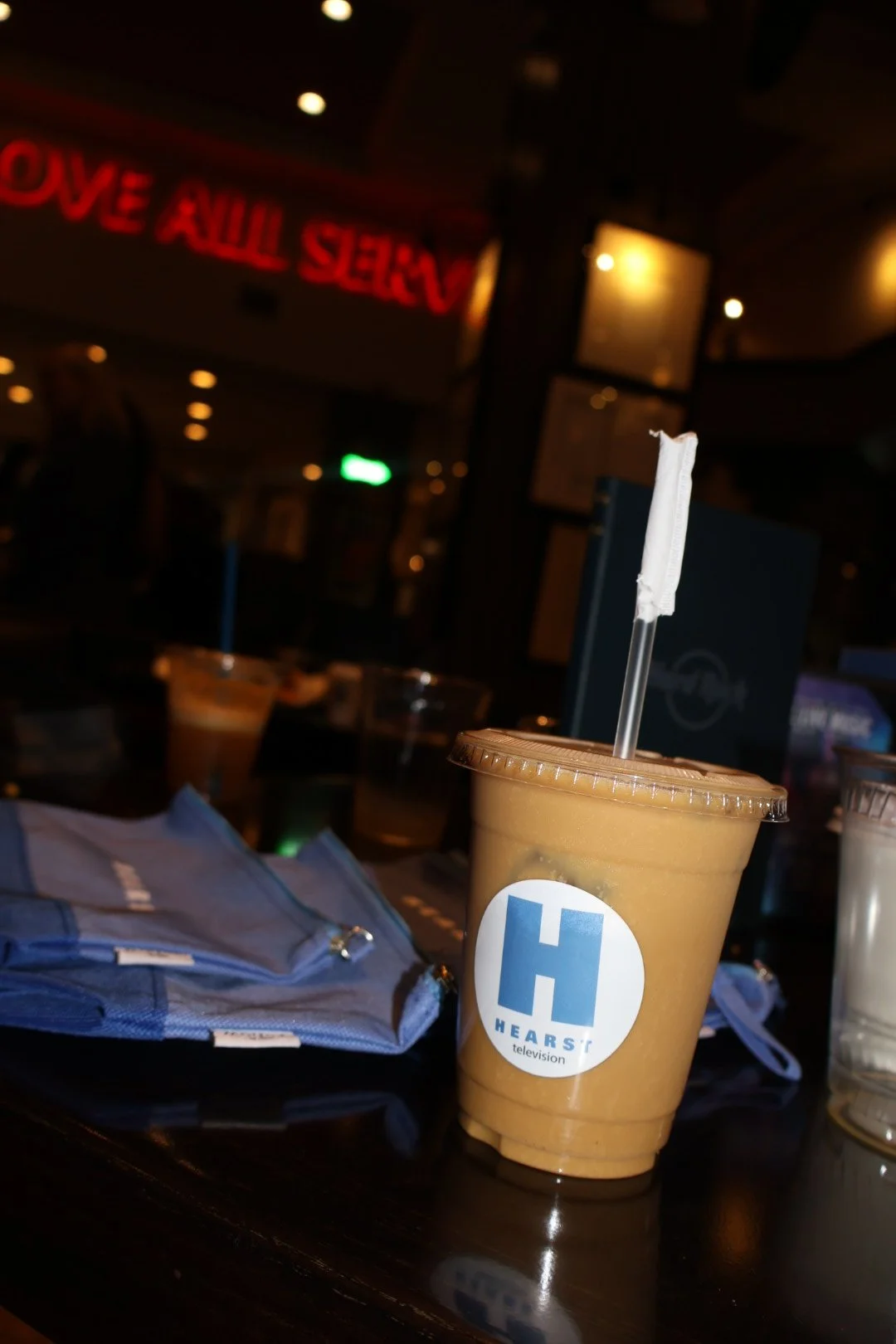 A table with a large iced coffee in a plastic cup with a straw, featuring a 'Hearst Television' logo, and some blue backpacks. In the background, a dimly lit indoor setting with a neon red sign and television screens.