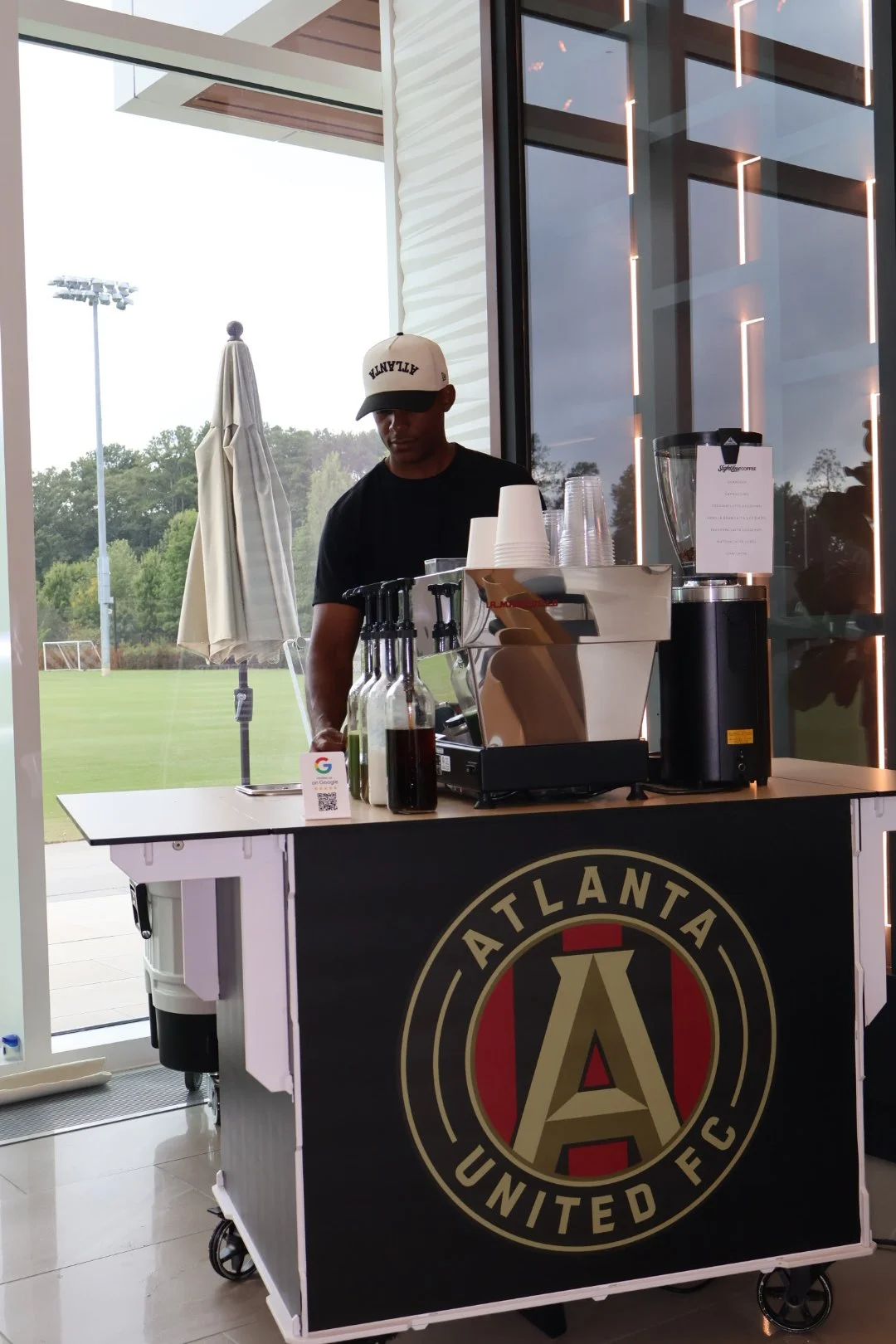 A man wearing a white cap with 'ATTITUDE' text stands behind a coffee cart with Atlanta United FC logo. The cart has coffee-making equipment, cups, and bottles, situated near a glass wall with a view of a grassy field and outdoor umbrella.