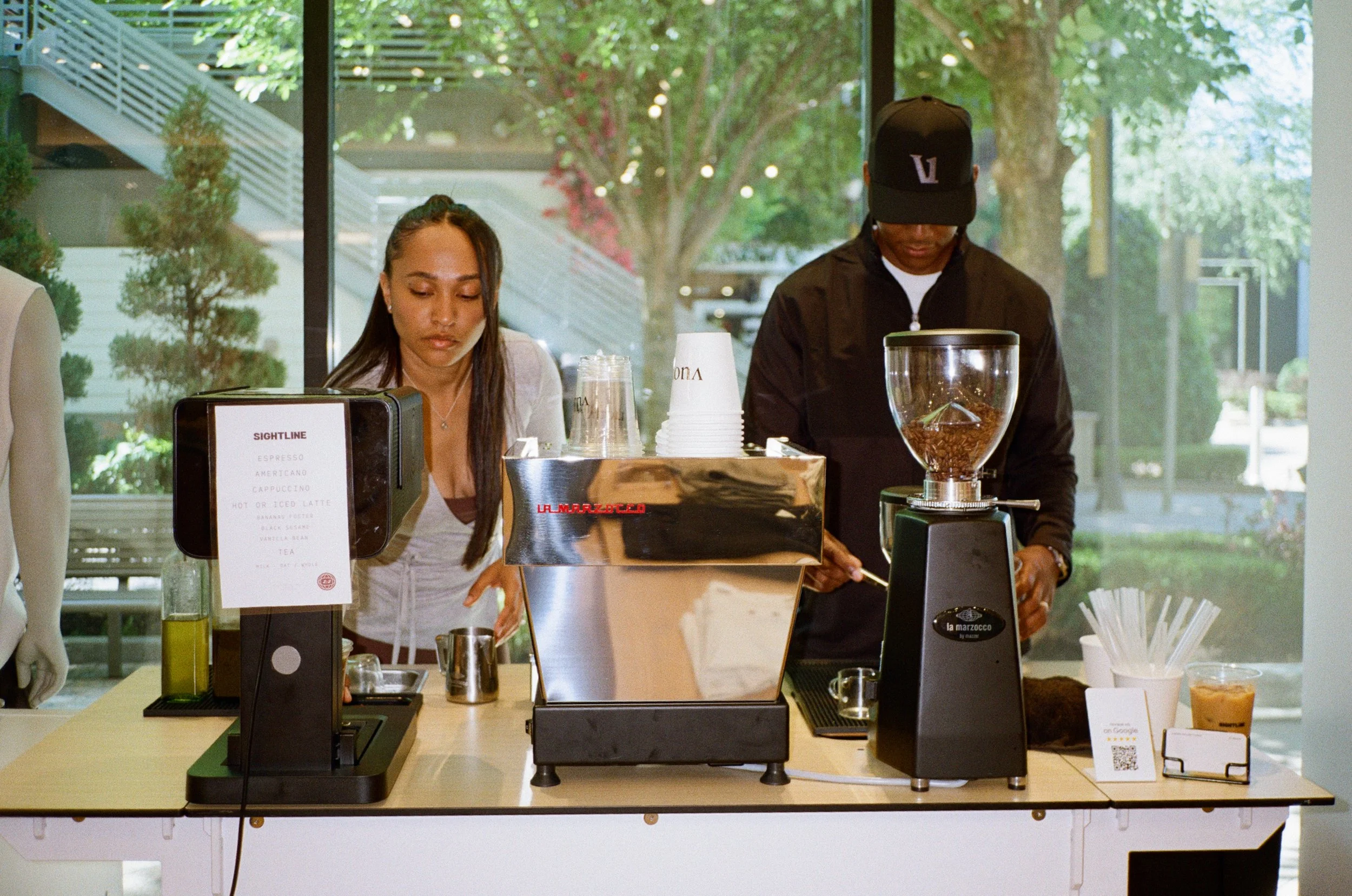 Two people behind a coffee counter, one woman and one man, preparing coffee with a coffee grinder, with a menu and cups visible in front of them, and a large window showing trees outside.
