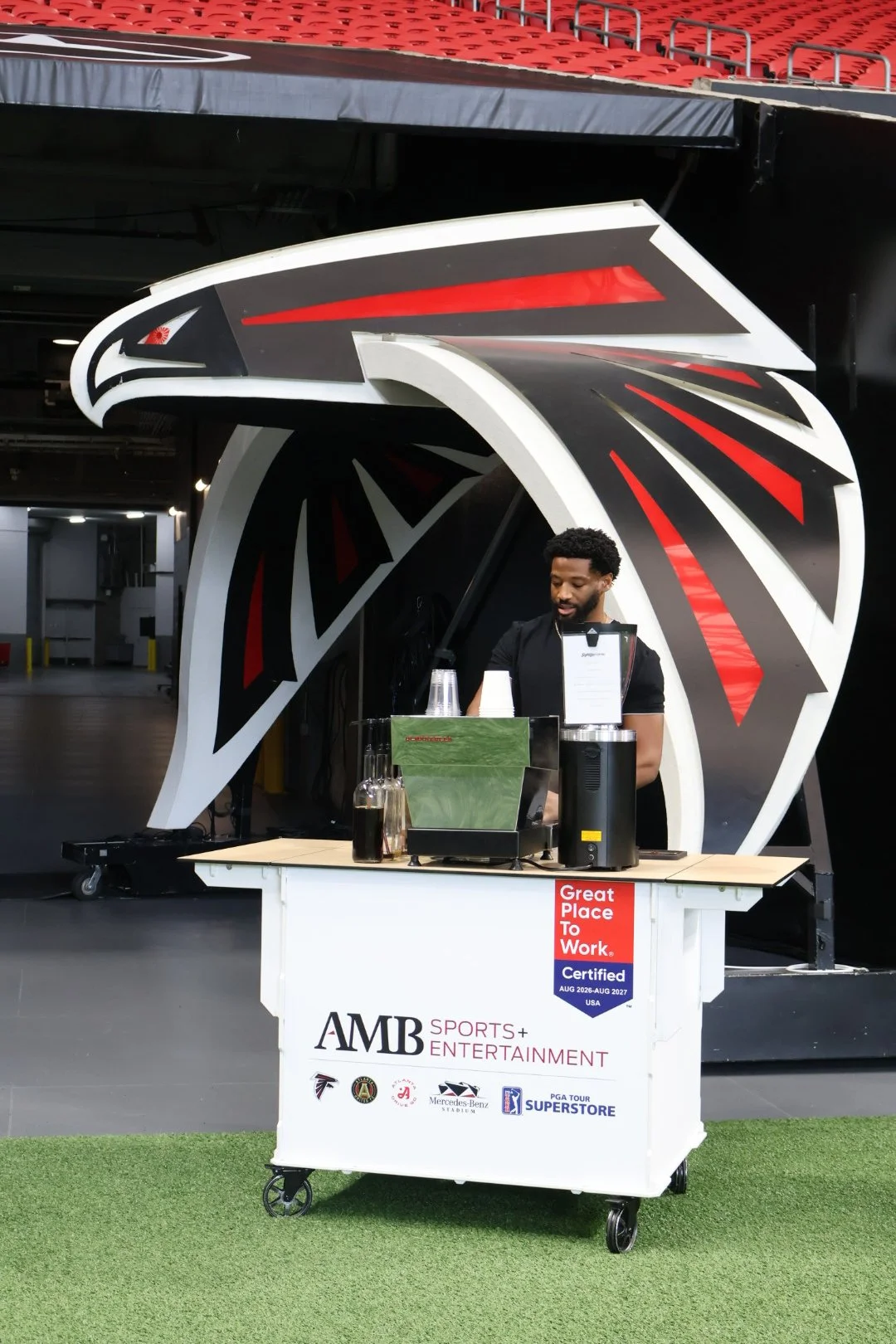 A drink stand with a Falcons football theme behind a man preparing beverages, with a sign that says 'Great Place To Work. Certified' and logos including Falcons, Mercedes-Benz Stadium, and PGA Tour Superstore.