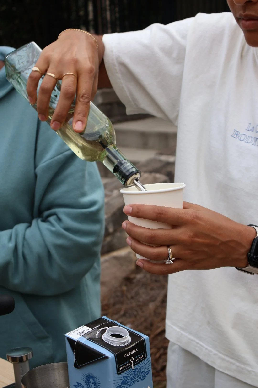 A person pouring a clear drink from a bottle into a cup held by another person, with a carton of oat milk nearby.