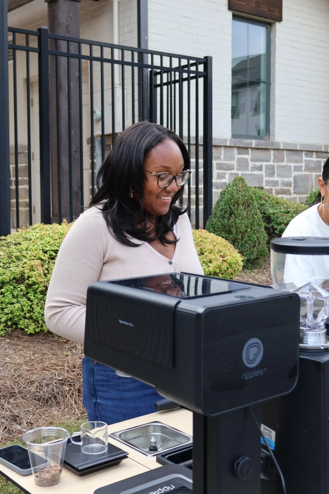 A woman with black hair and glasses smiling while standing next to an espresso machine on a custom coffee cart outdoors, with a house and greenery in the background.