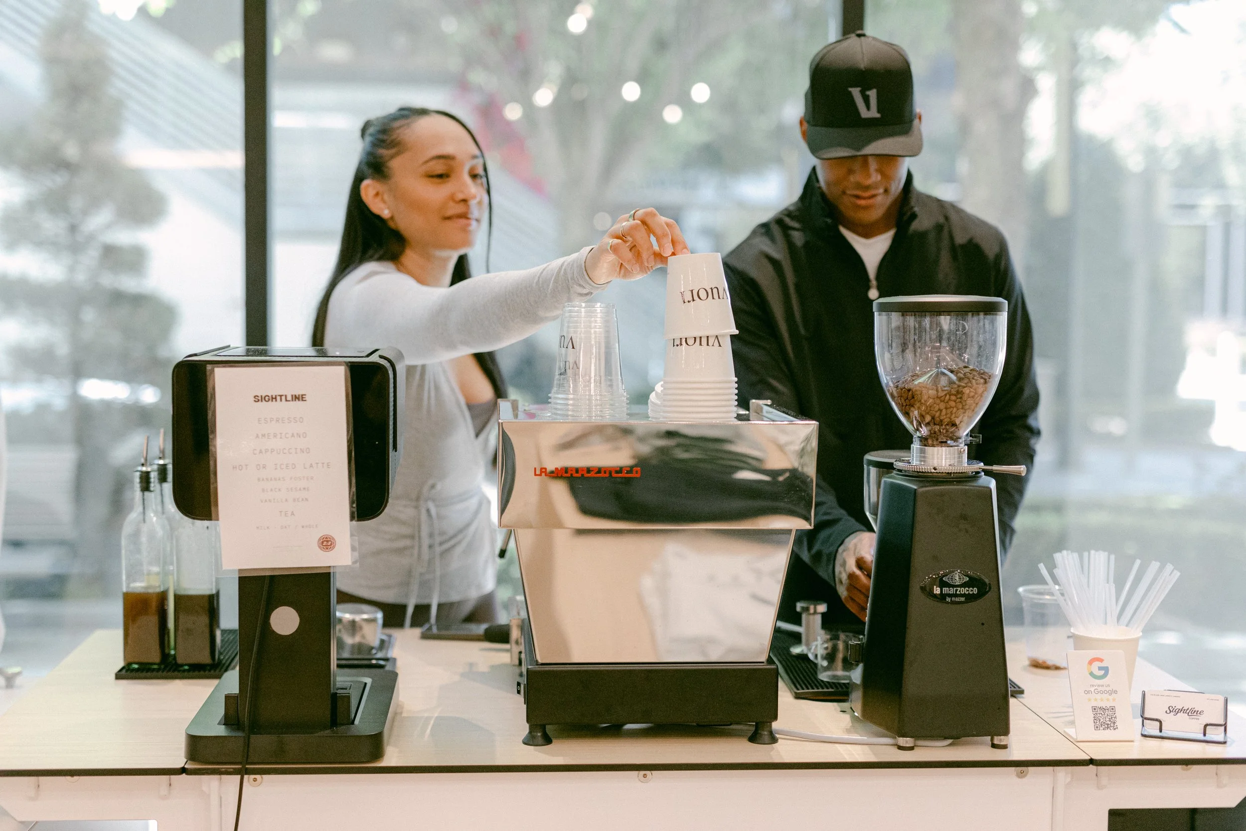 A woman and a man working together at a coffee stand, with the woman placing cups on the counter and the man using a coffee grinder, inside a well-lit cafe with large windows.