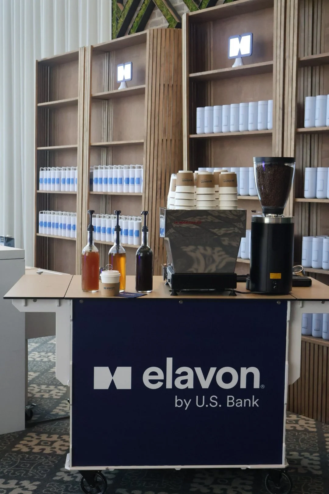 Coffee station with cups and bottled syrups, a coffee grinder, and a coffee machine, set against wooden shelves filled with white packages and a U.S. Bank branded table.