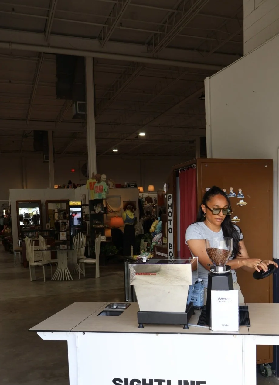 A woman with long black hair, wearing glasses, a gray t-shirt, and earrings, stands behind a coffee cart in a large retail store. Behind her are shelves with various items and a photo booth sign.