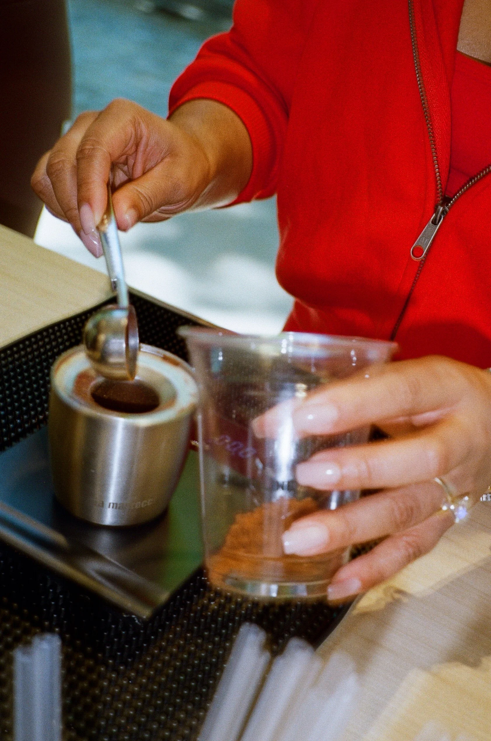Person in a red jacket holding a glass and using a spoon to serve ice cream from a metal container.