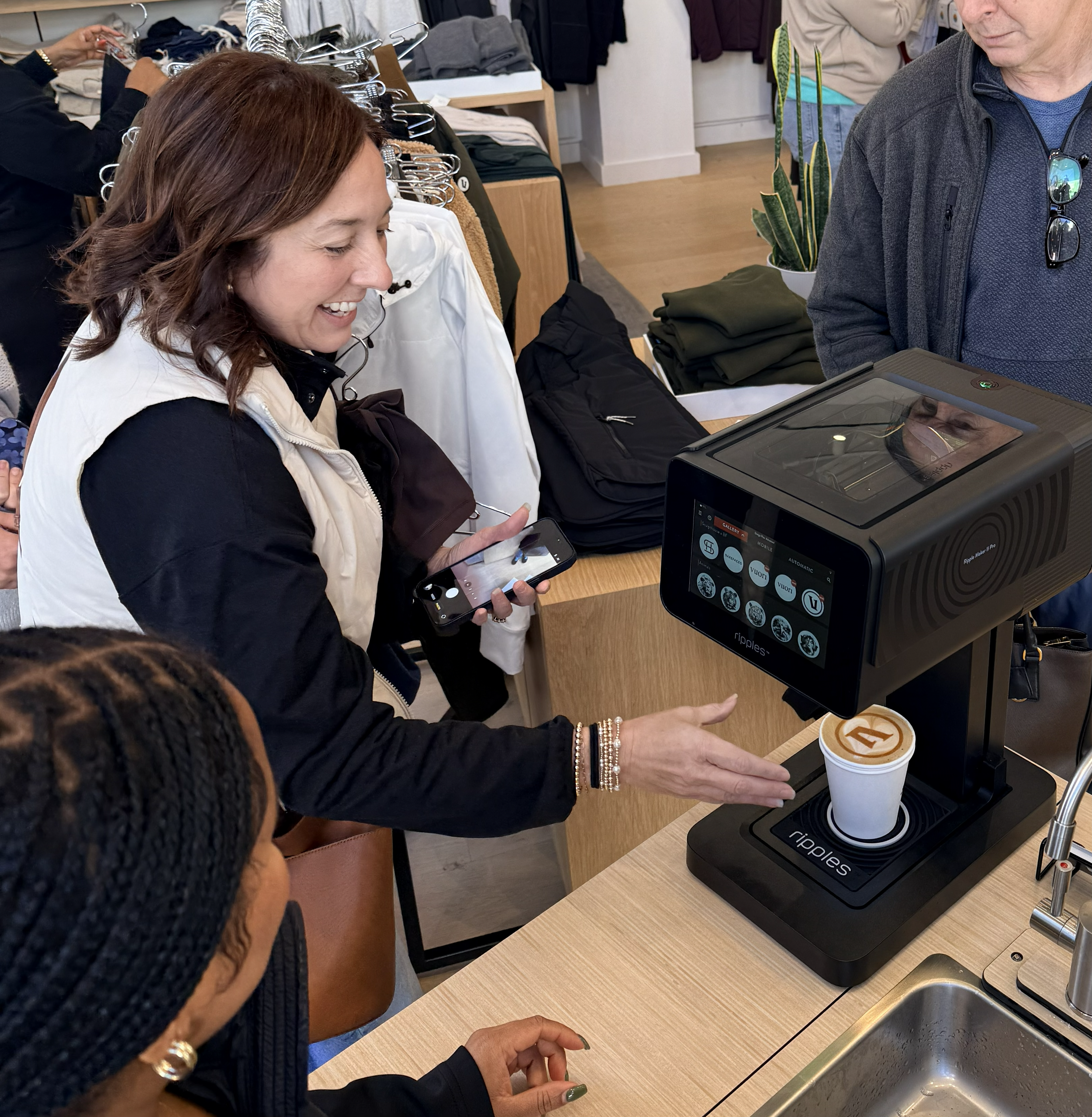 A woman at a store checkout counter is smiling and holding her phone, with a coffee cup on the counter nearby. There are other shoppers in the background, and a man standing next to her is looking at the screen of the device being used.