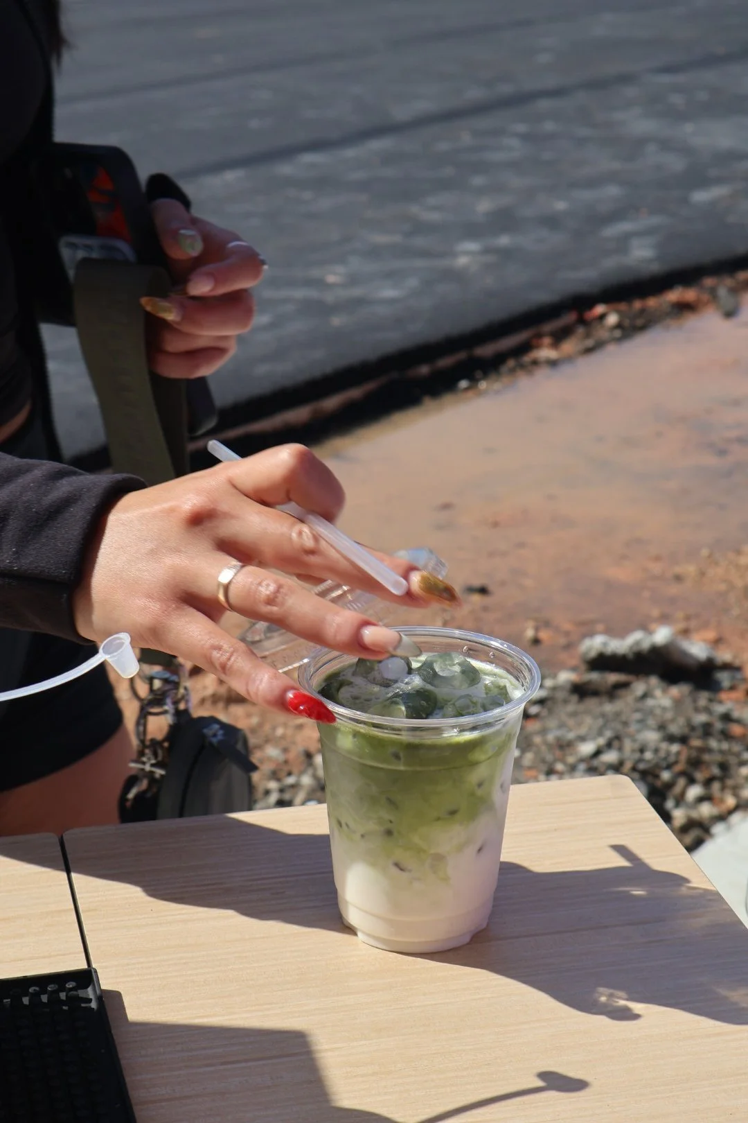 A person with long, decorated nails and a ring on their finger is mixing a matcha latte with ice in a clear plastic cup with a straw, outdoors on a wooden surface with a rocky background.