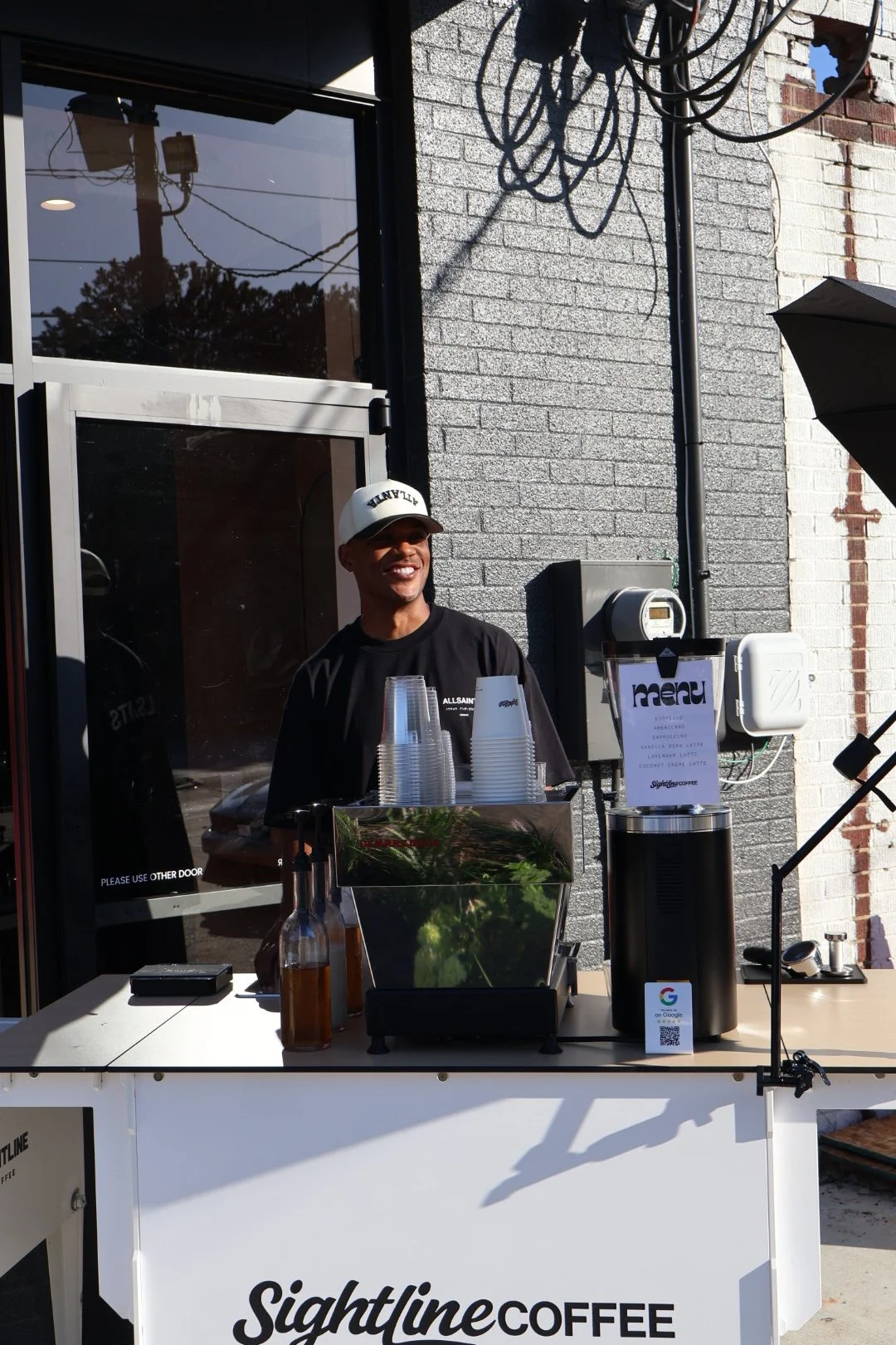 A smiling person wearing a white cap and black t-shirt stands behind a coffee cart with a sign that reads "Sightline Coffee." The cart has stacked cups, a bottle, and coffee brewing equipment, set against a brick building exterior with tangled black cables overhead.