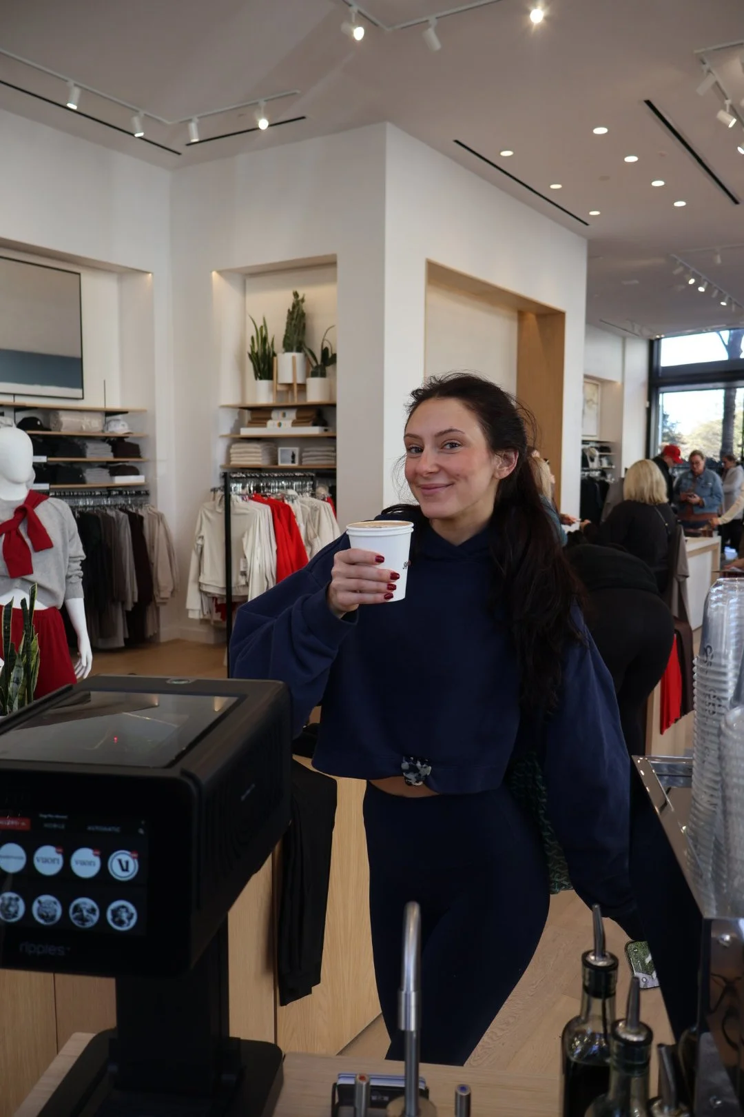 A woman standing behind a coffee bar smiling and holding a to-go coffee cup in a clothing store.