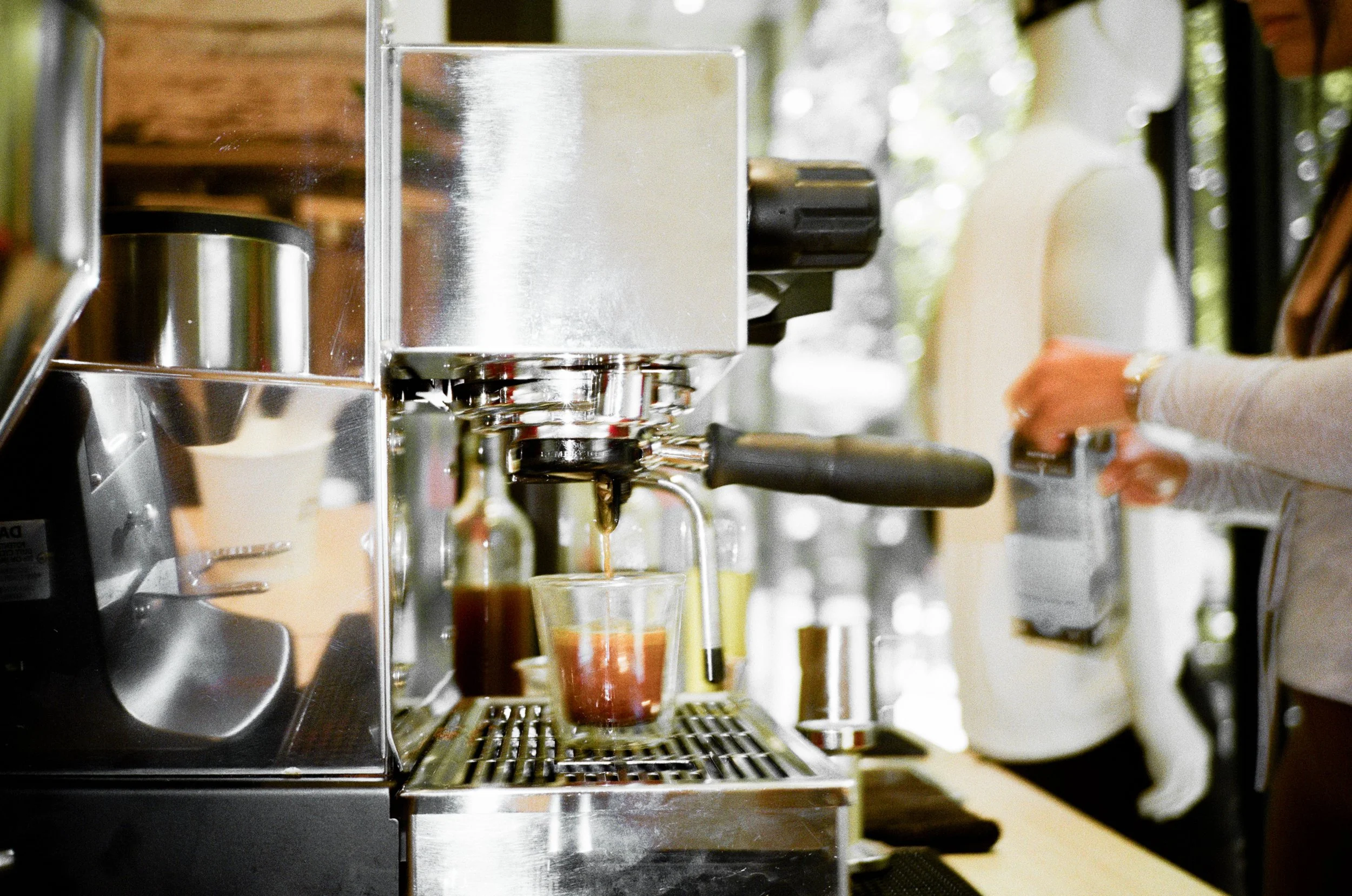 Close-up of espresso machine pouring coffee into a glass underneath the spout, with a person pressing a button in the background.
