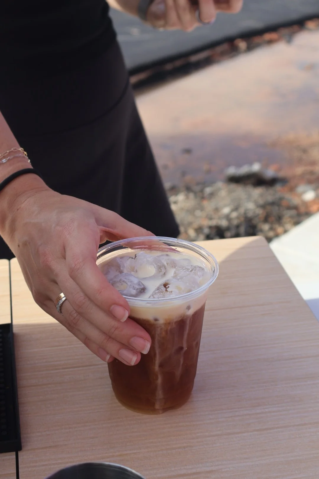 Close-up of a person's hand holding a plastic cup of iced coffee with milk on a wooden table, with a rocky beach and water in the background.