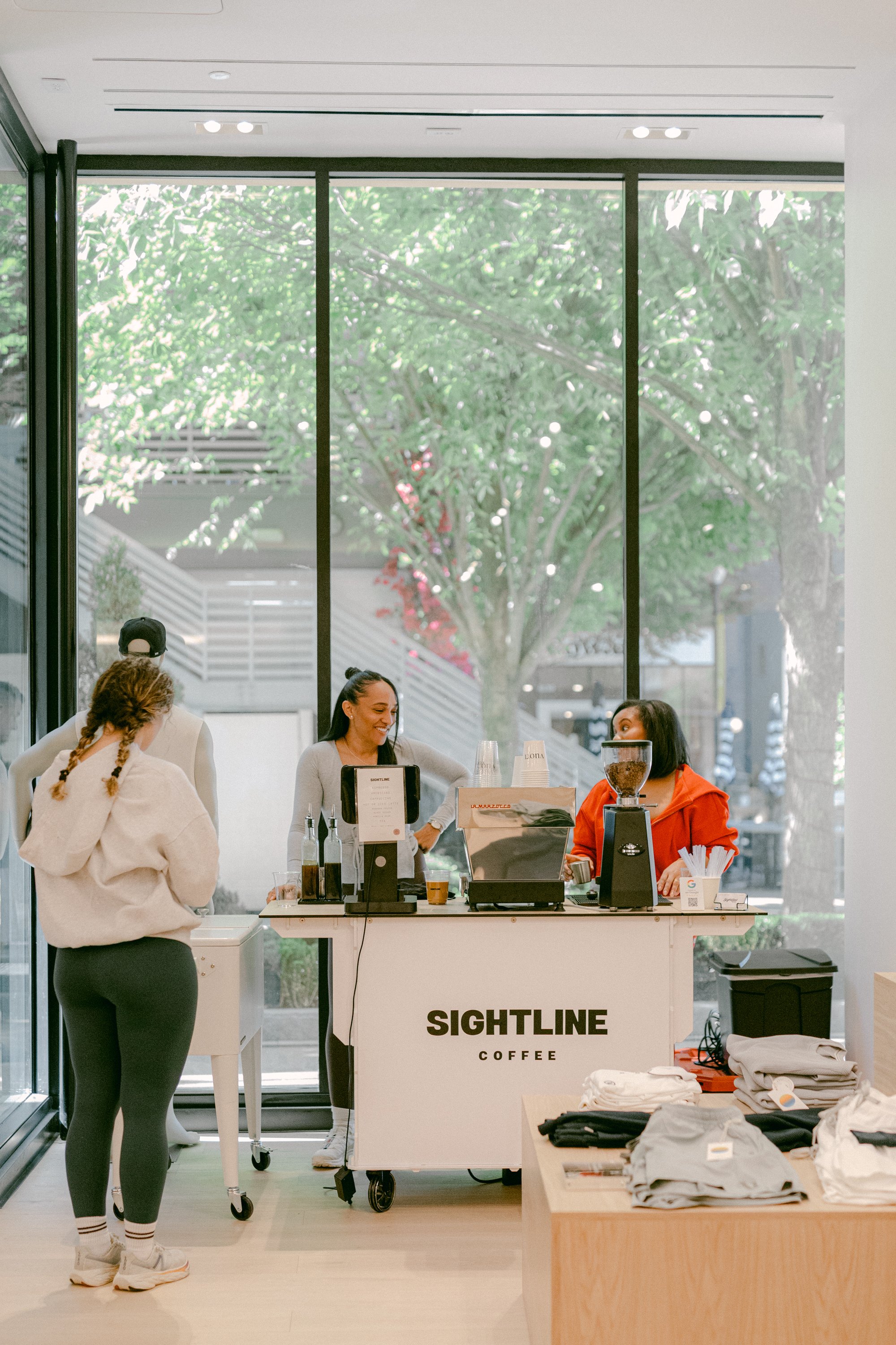 Customers ordering coffee at a counter inside a café with large windows and greenery outside, with branded signage reading 'SIGHTLINE COFFEE'.