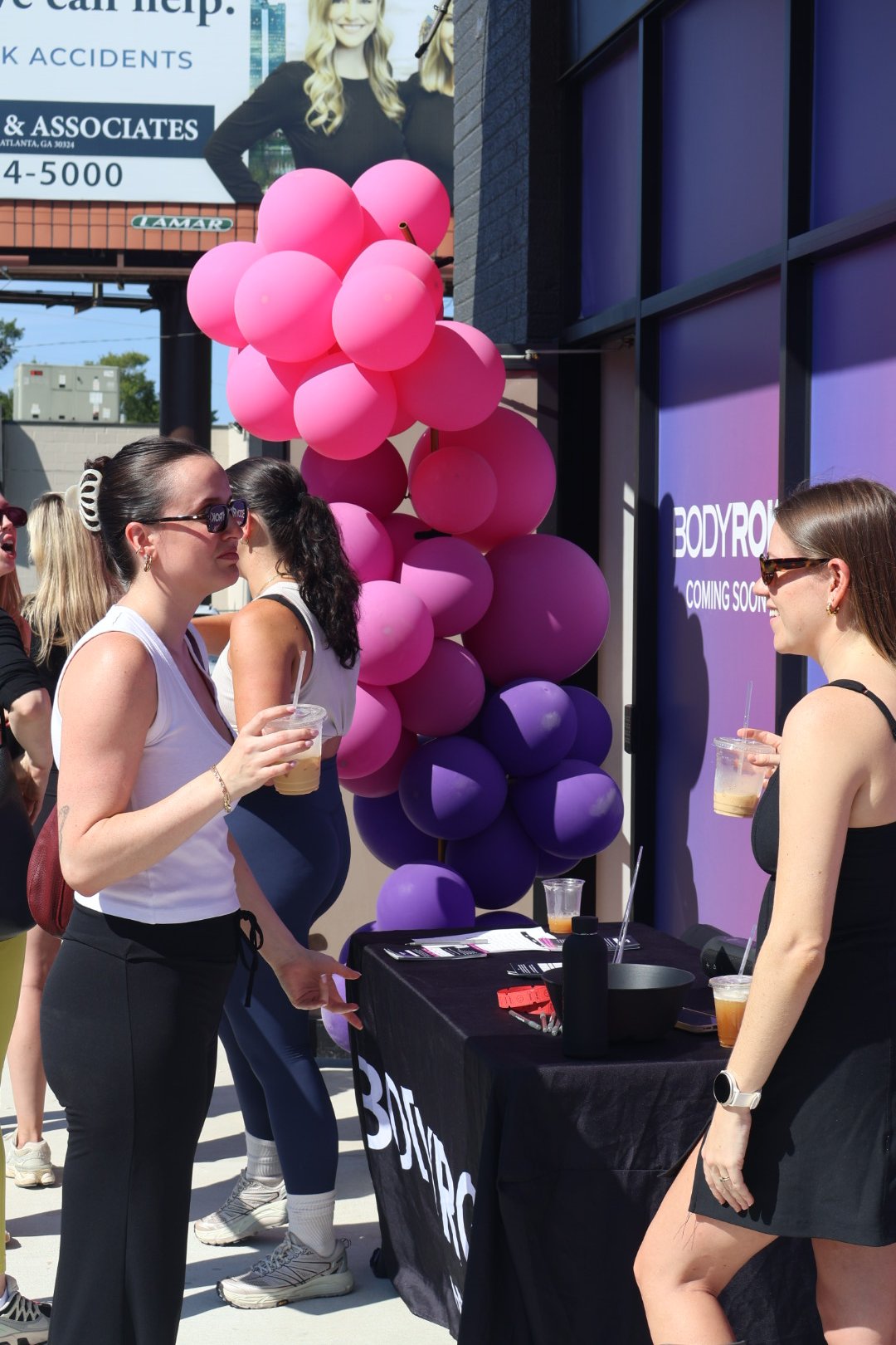Group of women at an outdoor event, standing near a table with a black tablecloth, drinks in hand, with pink and purple balloons in the background, and a purple sign that partially reads "BODY" and "COMING SOON."