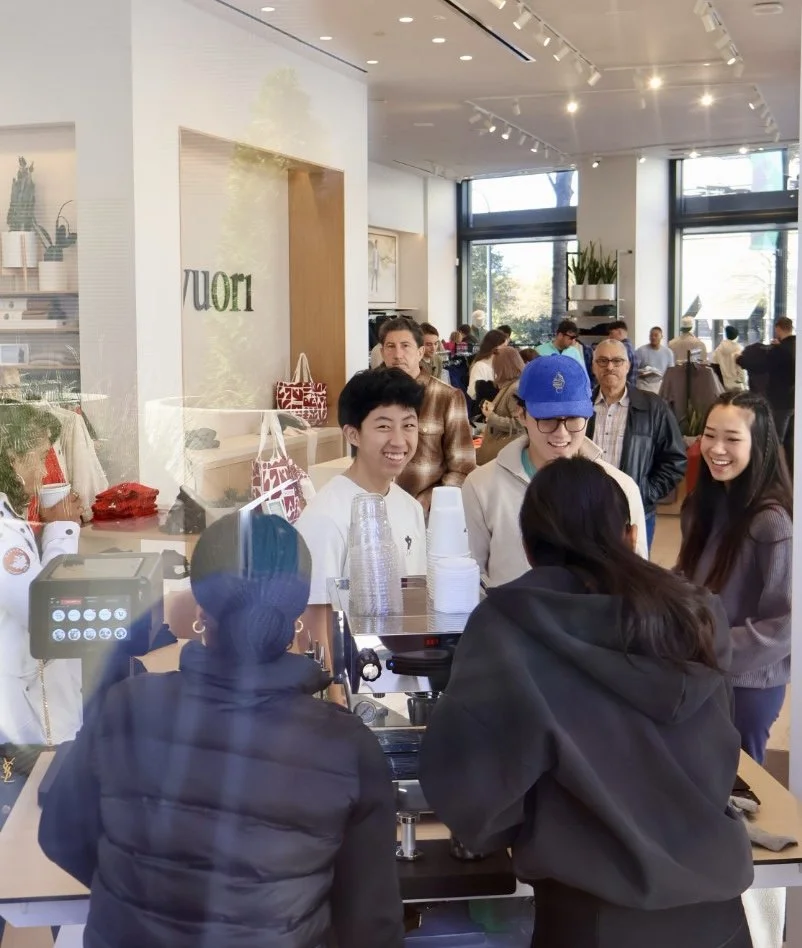 People smiling and ordering at a cafe counter in a modern, well-lit space with large windows, indoor plants, and artwork on the walls.