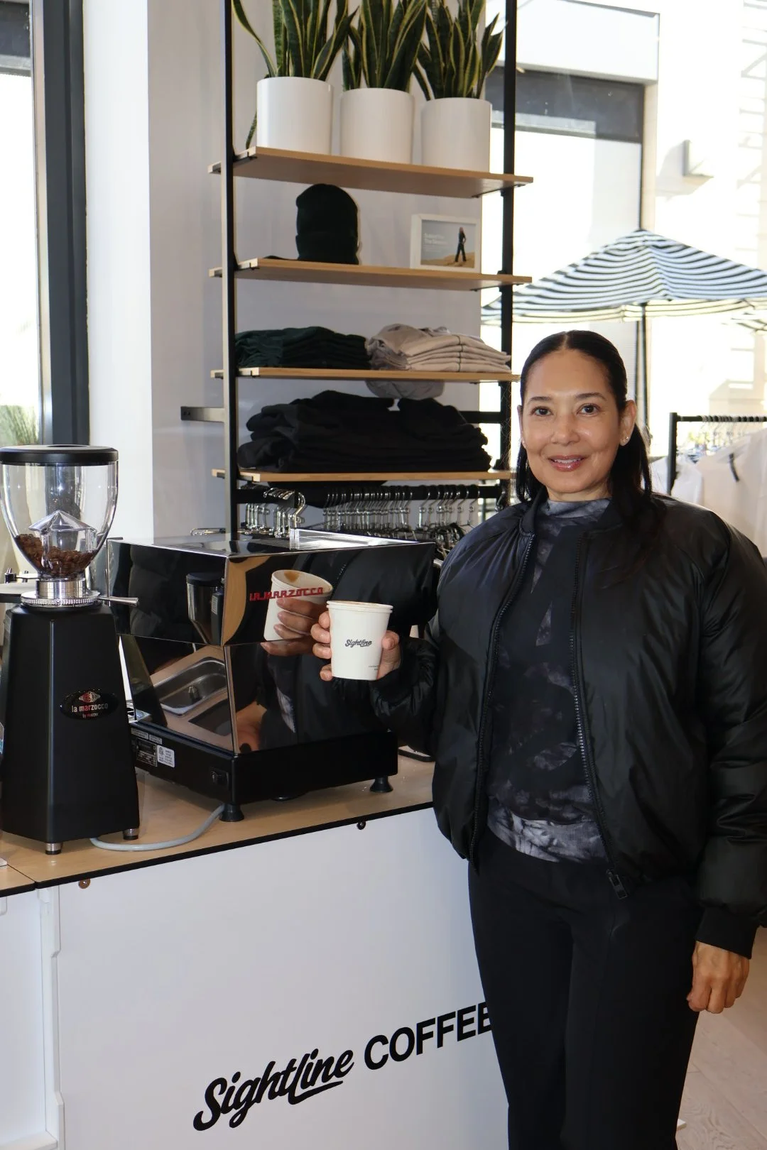 A woman with dark hair wearing a black jacket and dark pants, smiling while holding a cup of coffee in next to a custom coffee cart inside of a retail stoe.