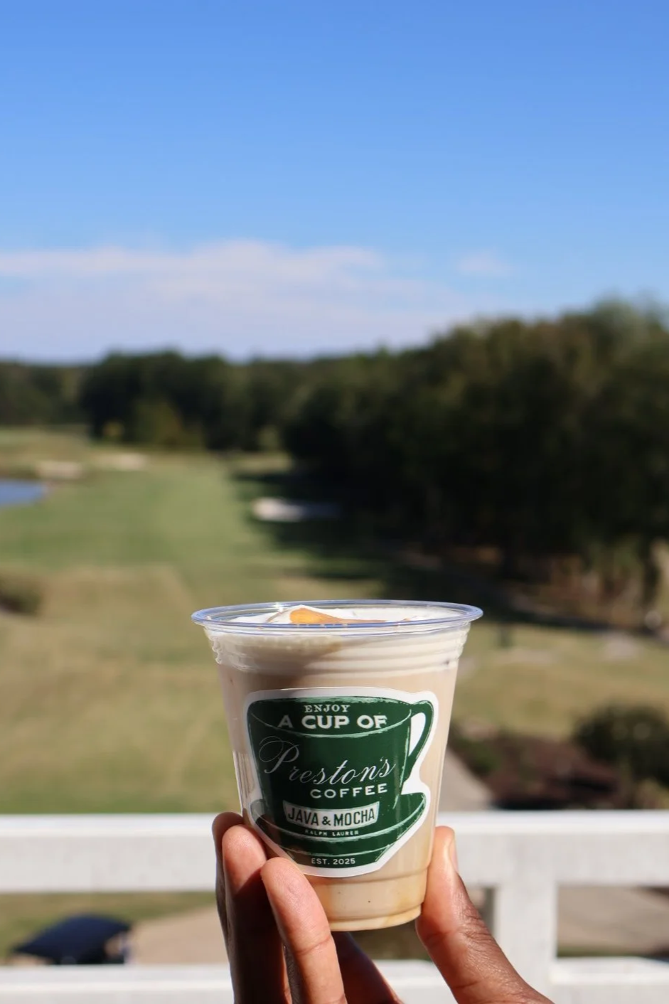 Hand holding a plastic cup with coffee labeled 'Preston's Coffee Java & Mocha' against a blurred outdoor landscape with trees and blue sky.