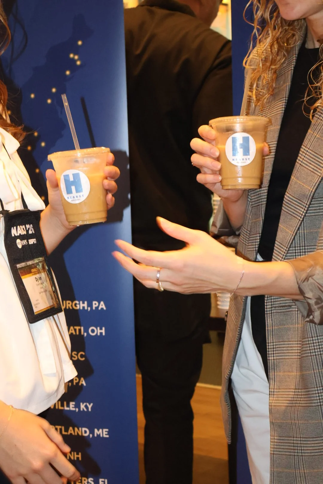 Two women holding iced coffee drinks with Harvard University logos, engaging in conversation at an event.