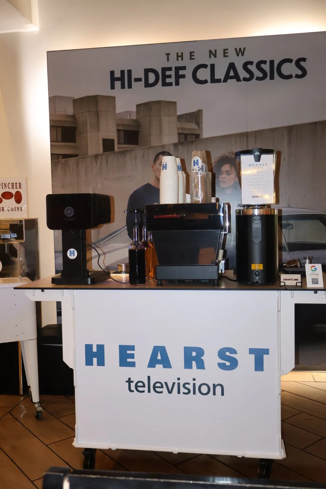 A self-serve coffee station with cups, a coffee machine, and a large black container in front of a banner advertising 'The New Hi-Def Classics'. The station has signage reading 'Hearst Television'.