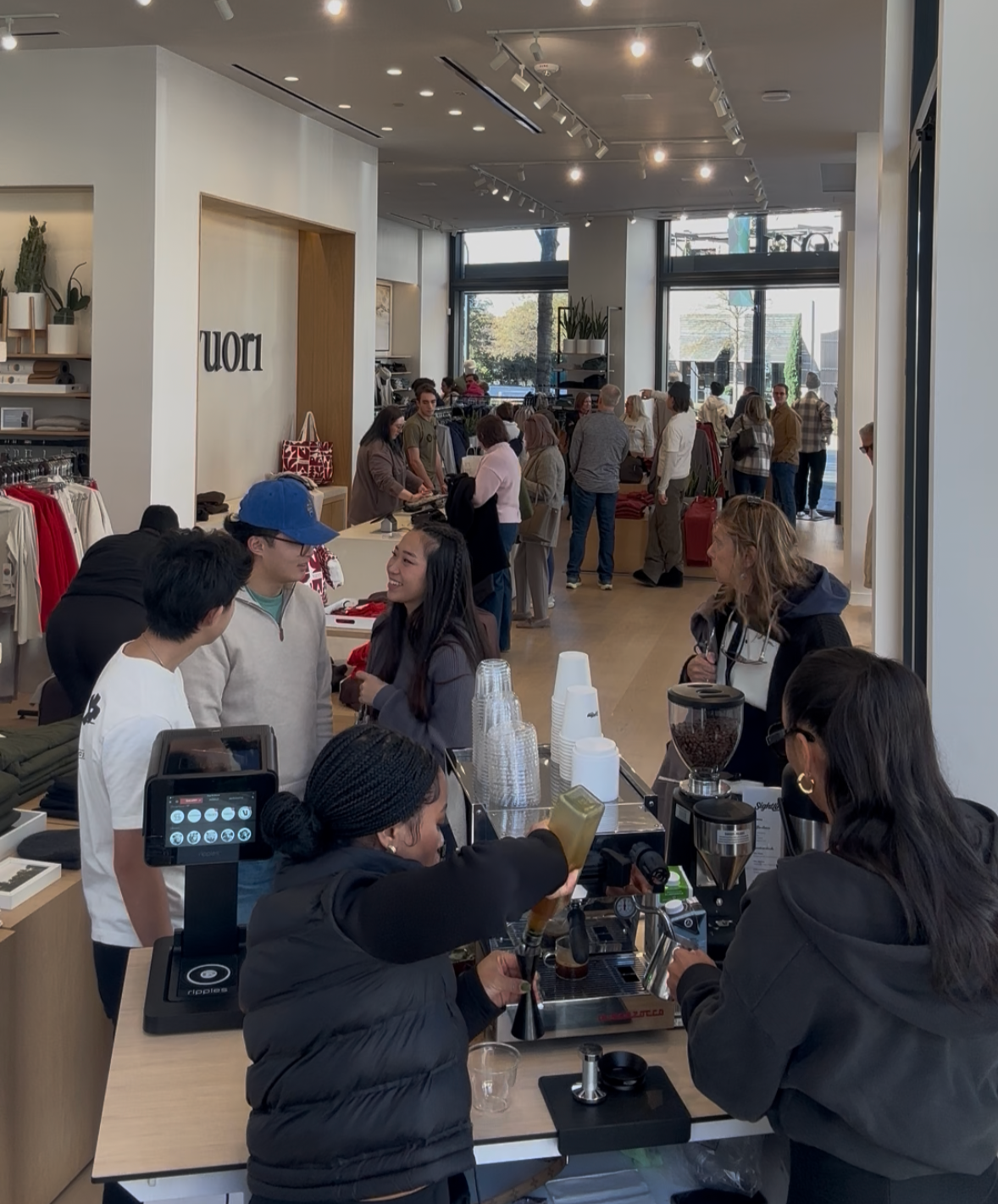 People shopping and ordering coffee at a retail store, with a busy checkout counter in the foreground and racks of clothes and customers browsing in the background.