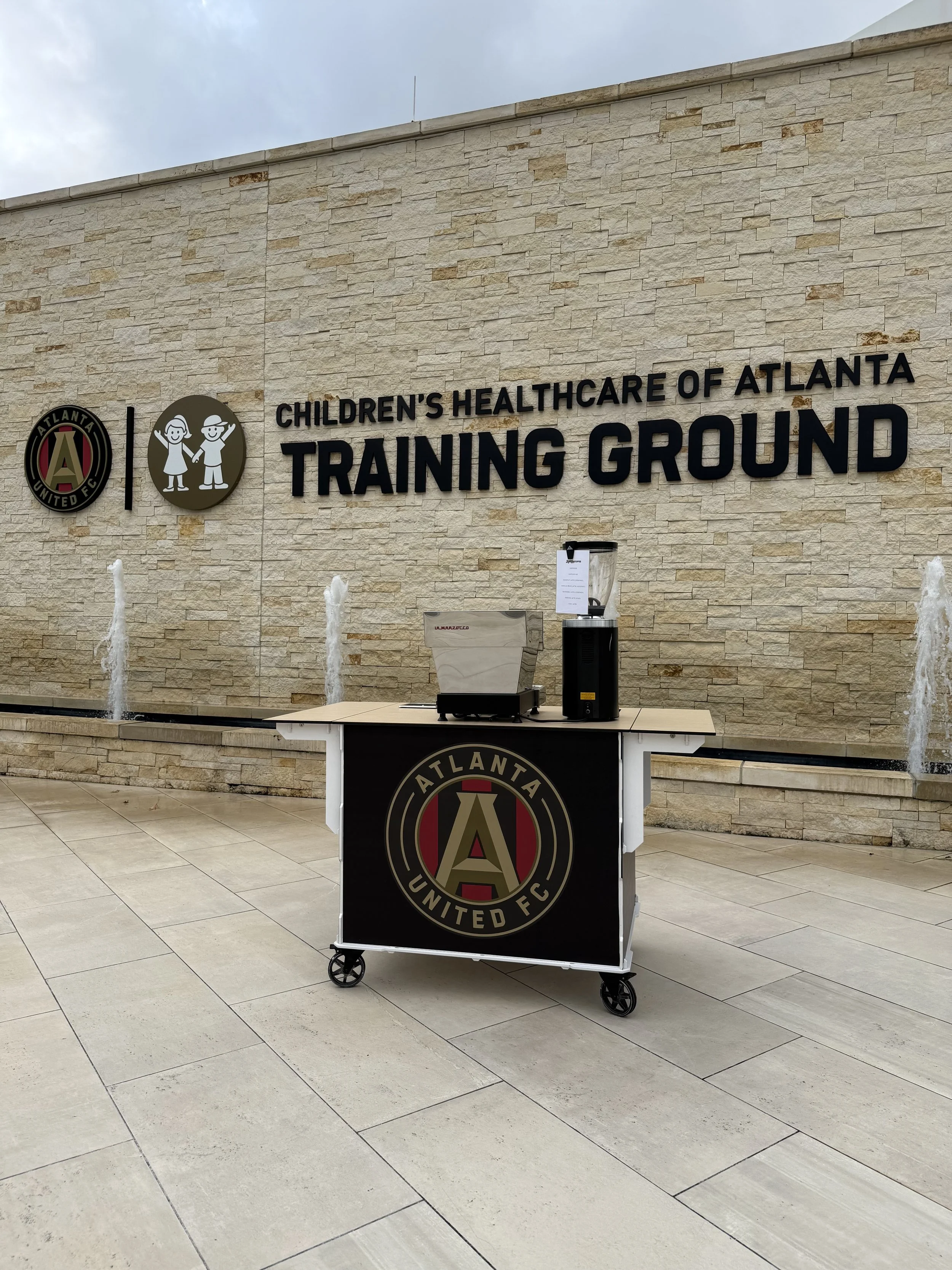 A beige brick wall with black lettering reading 'Children's Healthcare of Atlanta Training Ground' and logos of Atlanta United FC, a black table on wheels with a black curtain and a water cooler in front of it, with a tiled floor and water fountains on each side.