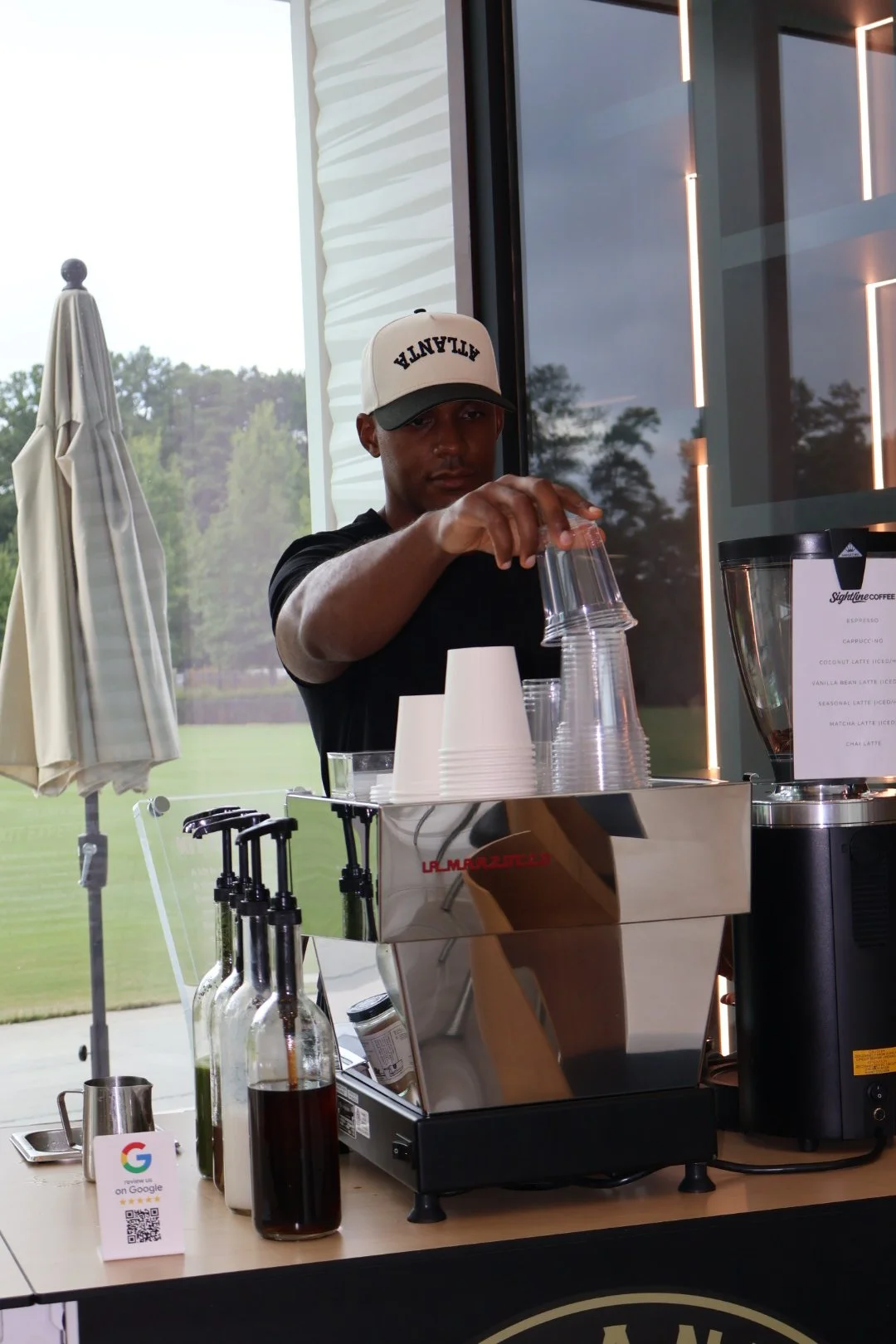 A person making coffee at a counter with a commercial espresso machine, coffee syrups, plastic cups, and a menu, inside a cafe or coffee shop with large windows showing trees outside.