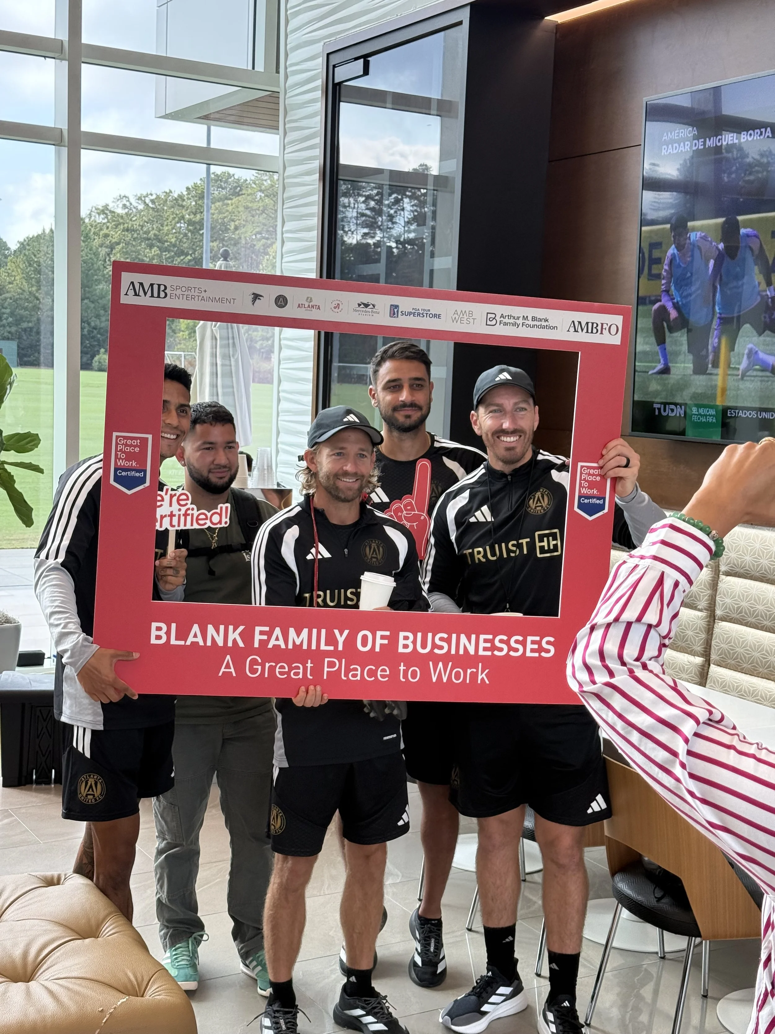Group of five men in black Adidas sportswear holding a red sign with white text, standing inside a building near a window and a large screen showing a soccer match. Some are smiling, one has a beard and long hair, and one is wearing a cap. The sign reads, 'A Great Place to Work!'.