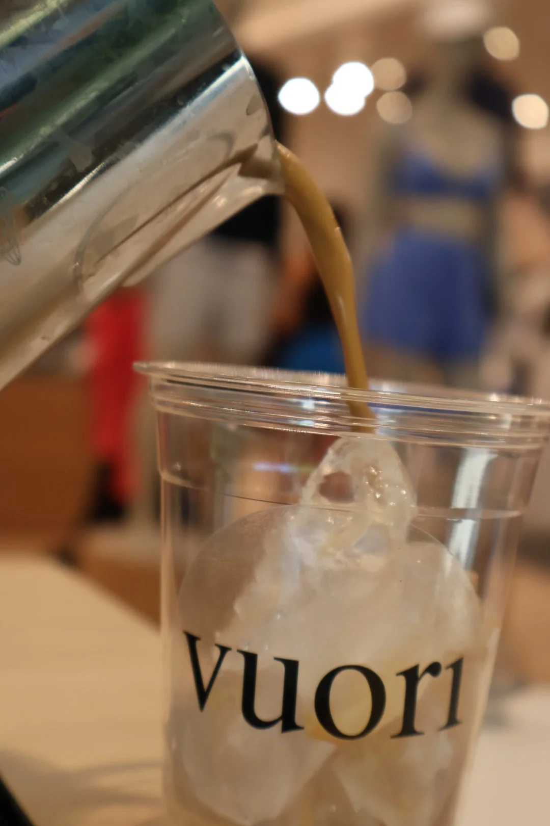 A beverage being poured from a can into a plastic cup with ice cubes, in a setting that appears to be a cafe or restaurant.