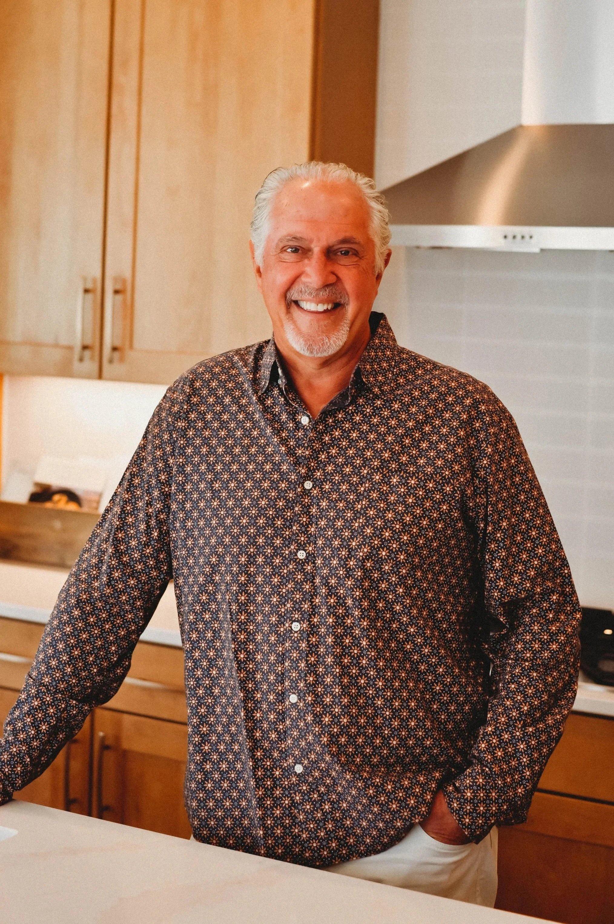 A smiling older man with gray hair and a beard, wearing a patterned button-up shirt, standing in a modern kitchen.