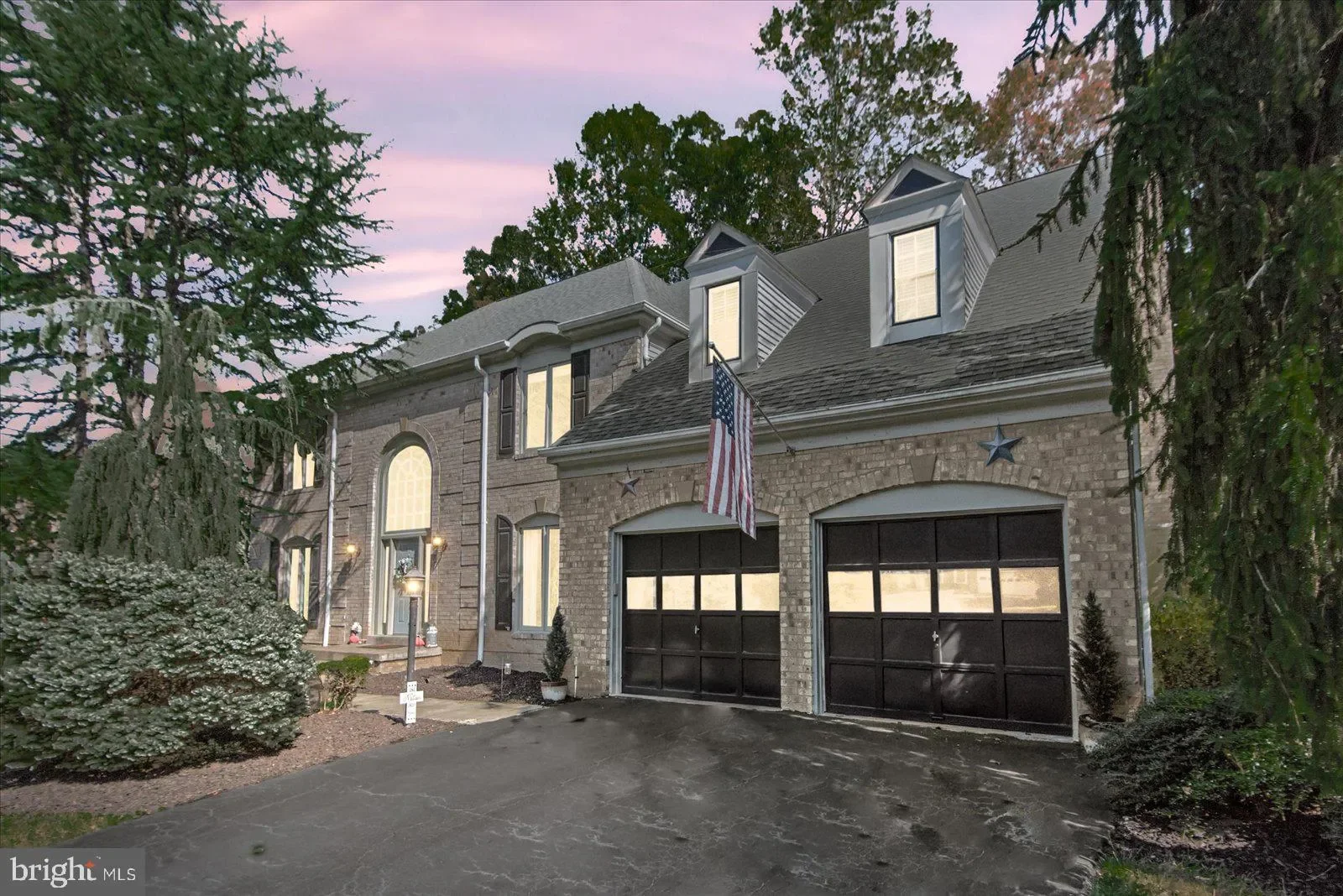 A two-story house with brick exterior, black garage doors, and dormer windows, decorated with an American flag and star accents, during dusk with trees and a pinkish-purple sky in the background.