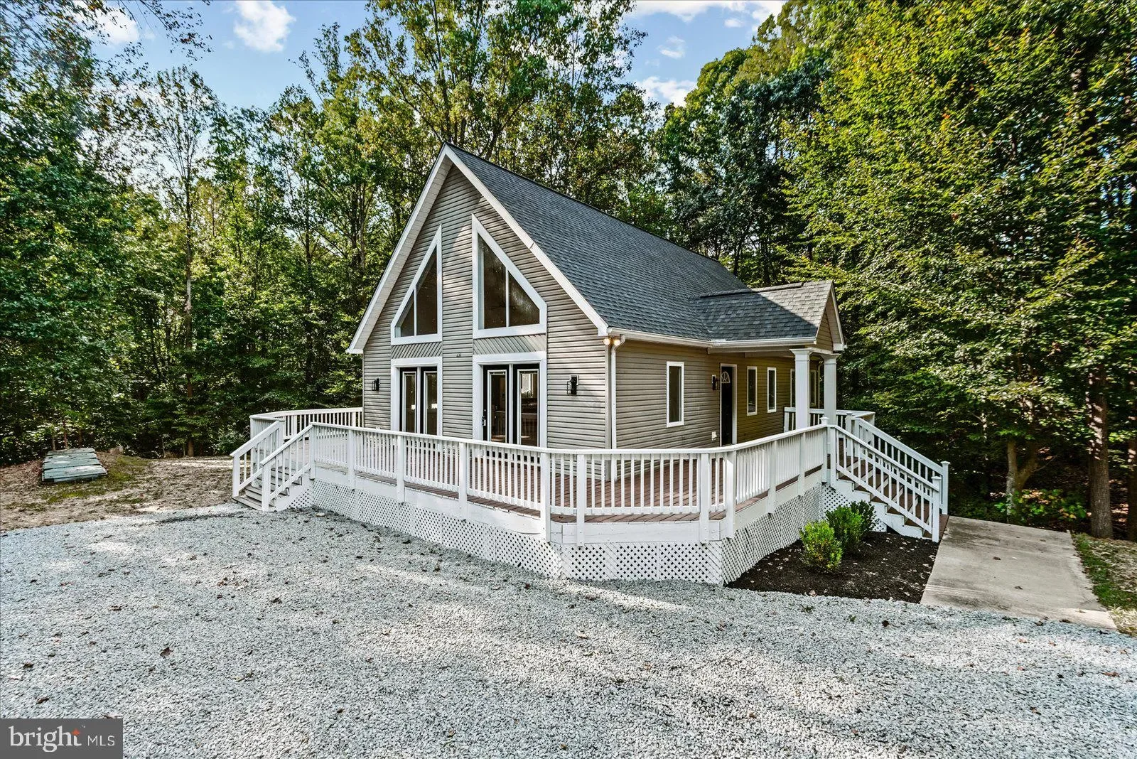 A two-story house with gray siding, a steep gabled roof, and large triangular windows on the upper floor. It has a wraparound concrete deck with white railing, surrounded by trees.