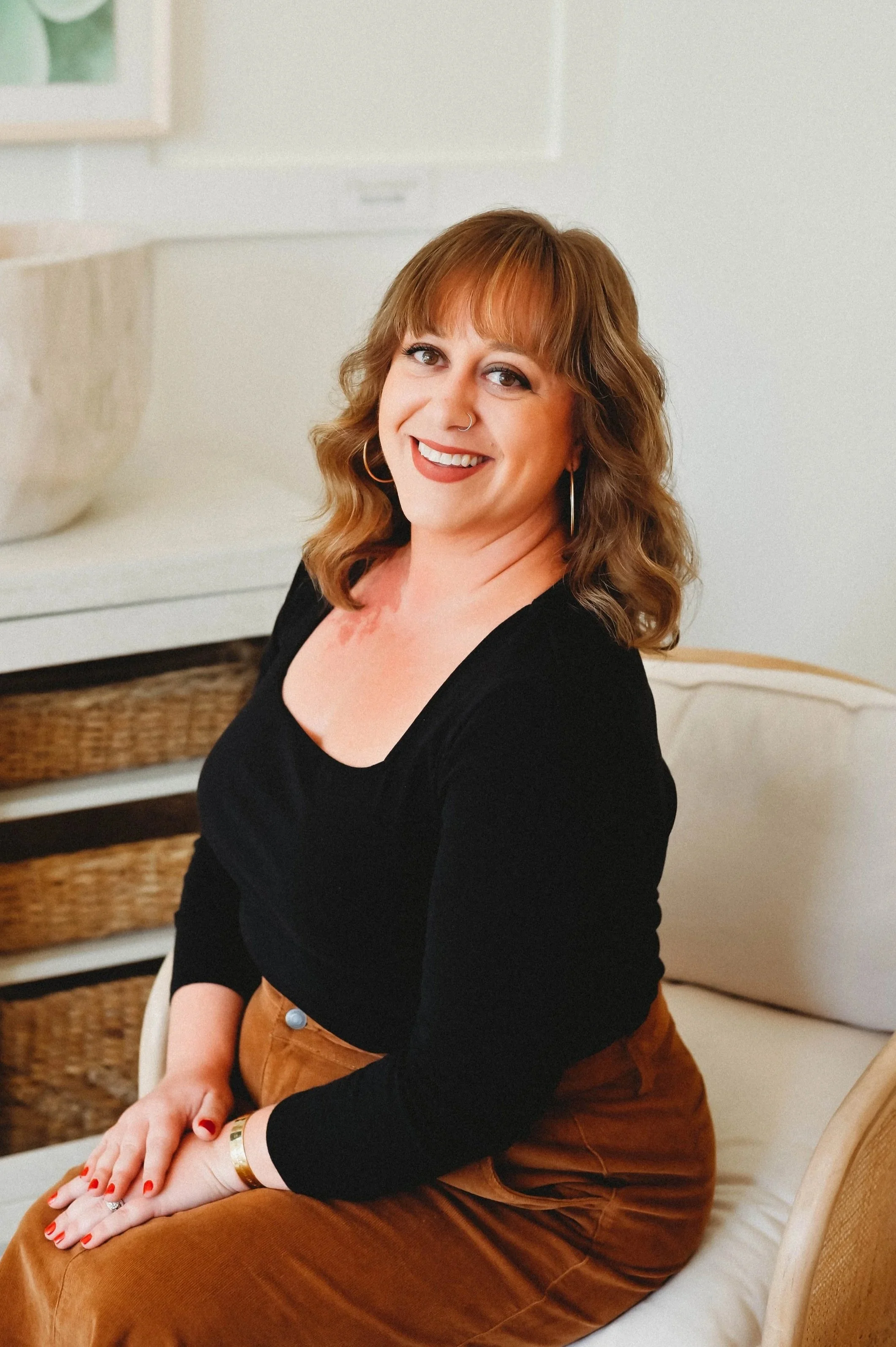 A woman with shoulder-length wavy hair, wearing a black top and brown pants, sitting on a beige chair, smiling at the camera, in a room with neutral decor.