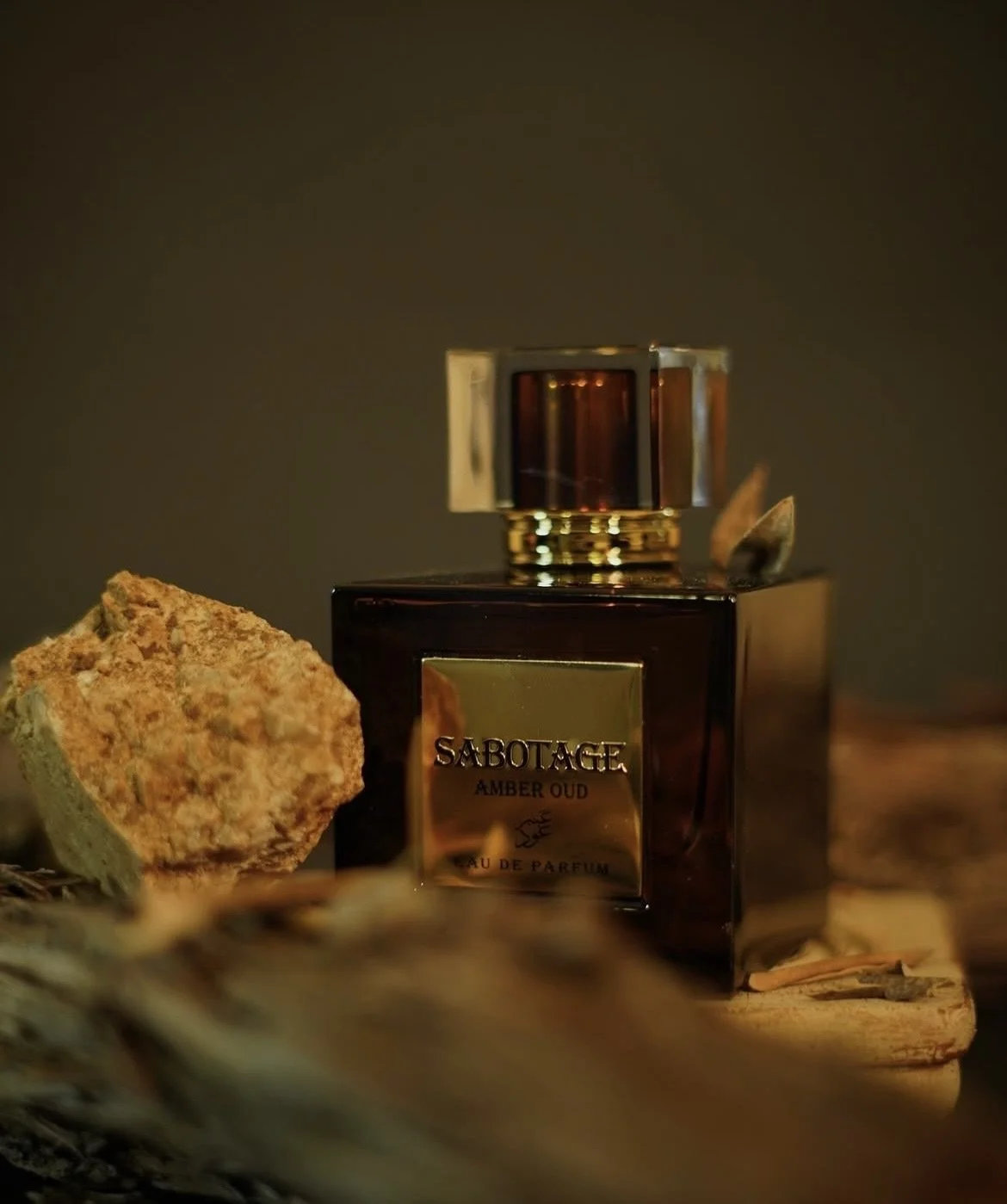 A bottle of Sabotage Amber Oud perfume surrounded by rocks and driftwood, set against a dark background.