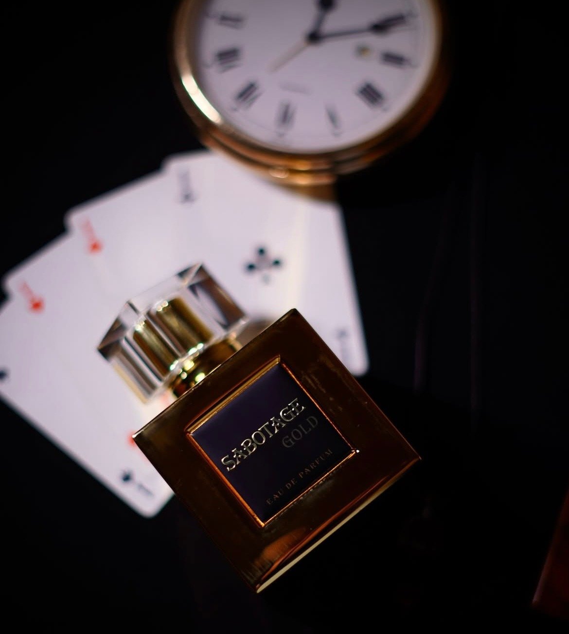 A bottle of Sabotage Gold perfume, a wristwatch, playing cards, and poker chips on a dark surface.